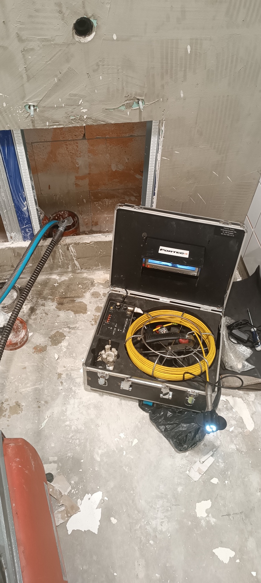 Kamera inspekcyjna w metalowej walizce obok otworu w ścianie z wystającą rurą. Sprzęt do inspekcji kanalizacji i udrażniania rur.