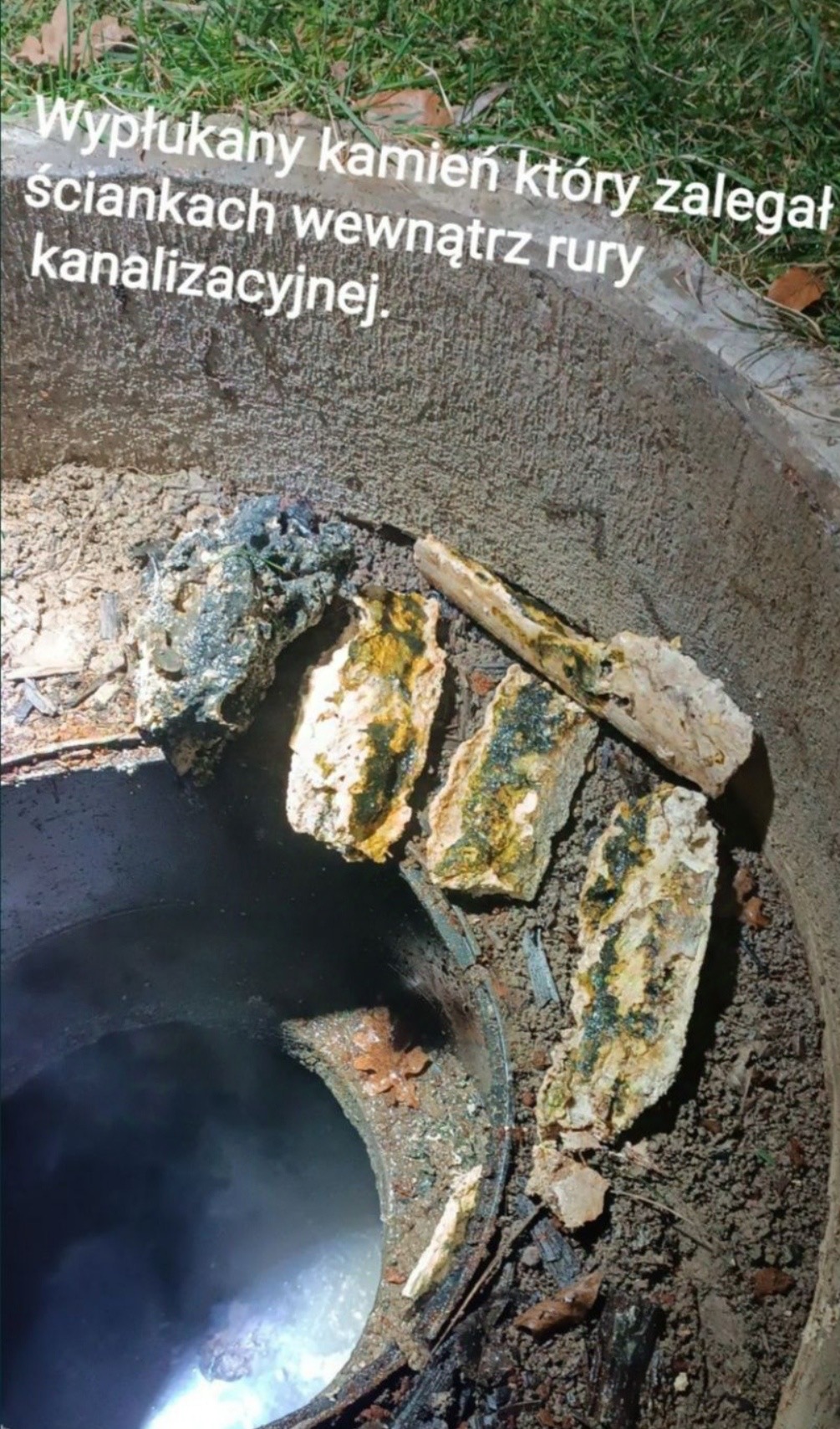 Otwarty właz kanalizacyjny z widocznymi kamieniami i osadami usuniętymi z wnętrza rury. Widoczne zanieczyszczenia na betonie i resztki ziemi.