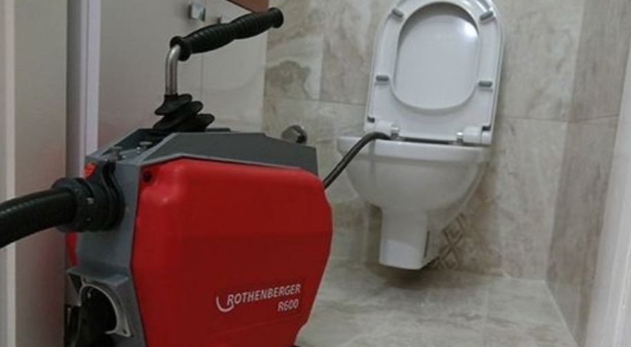 Toaleta z otwartą klapą i urządzenie Rothenberger R600 do udrażniania rur, z widoczną giętką rurą wewnątrz muszli, na tle beżowych płytek.