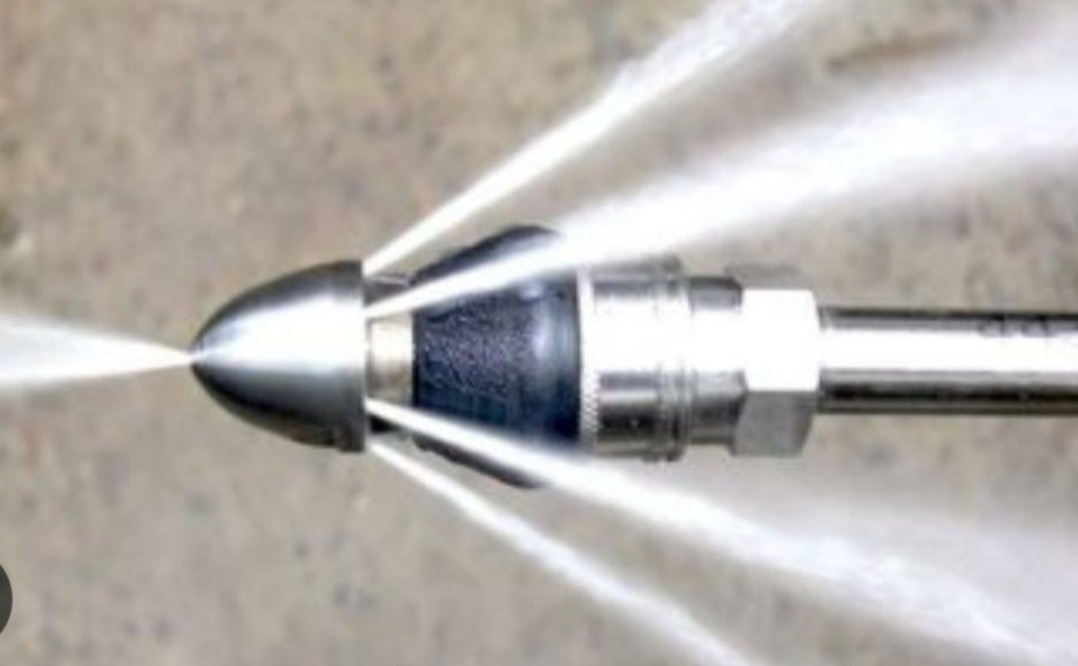 Dysza udrażniająca rury pod ciśnieniem, woda tryska z otworów, metalowa konstrukcja, szary beton w tle. Widok z bliska.