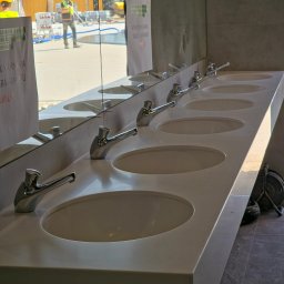 BISHORN SERGII KRAVCHENKO - Długa, biała umywalka z kilkoma kranami, odbicie w lustrze pokazuje budowę na zewnątrz. Umywalka o minimalistycznym designie, z widocznymi znakami ostrzegawczymi.