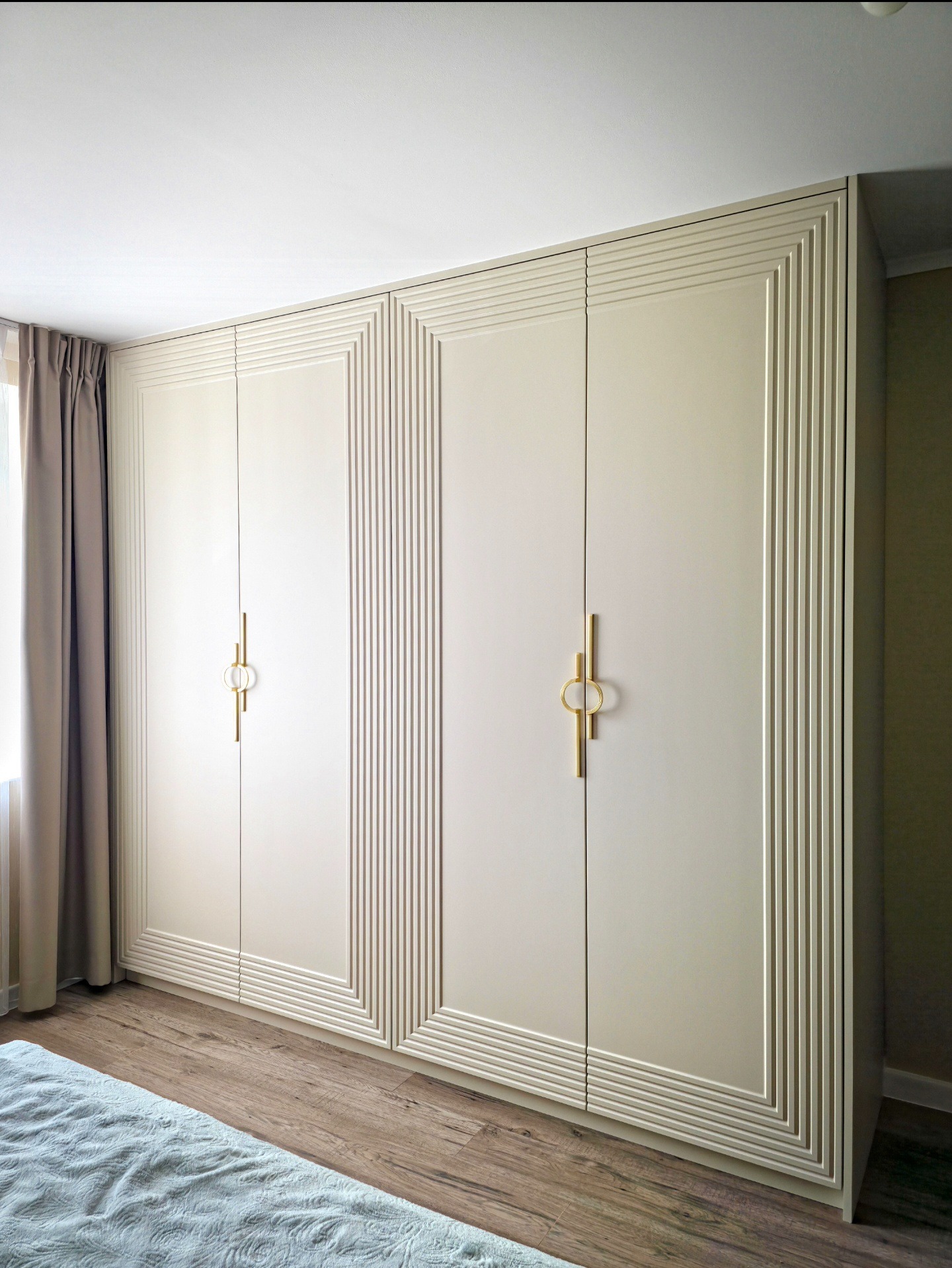 Elegancka, beżowa szafa na wymiar z dekoracyjnymi frezowaniami i złotymi uchwytami, dopasowana do wnętrza sypialni z drewnianą podłogą i zasłonami.