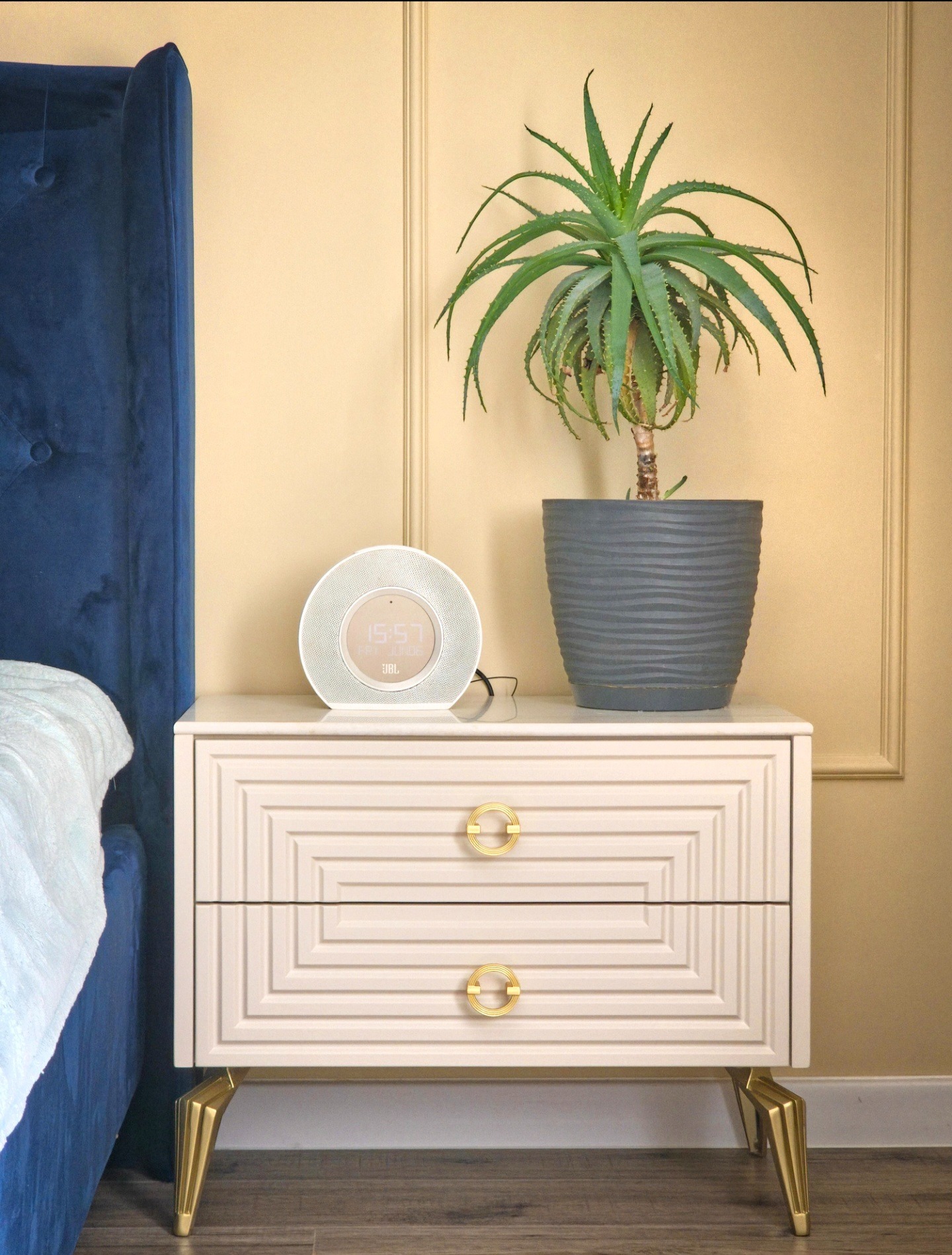 Beżowa szafka nocna z dwoma szufladami, złotymi uchwytami i nóżkami, obok niebieskiego łóżka. Na szafce roślina w szarej doniczce i białe radio.