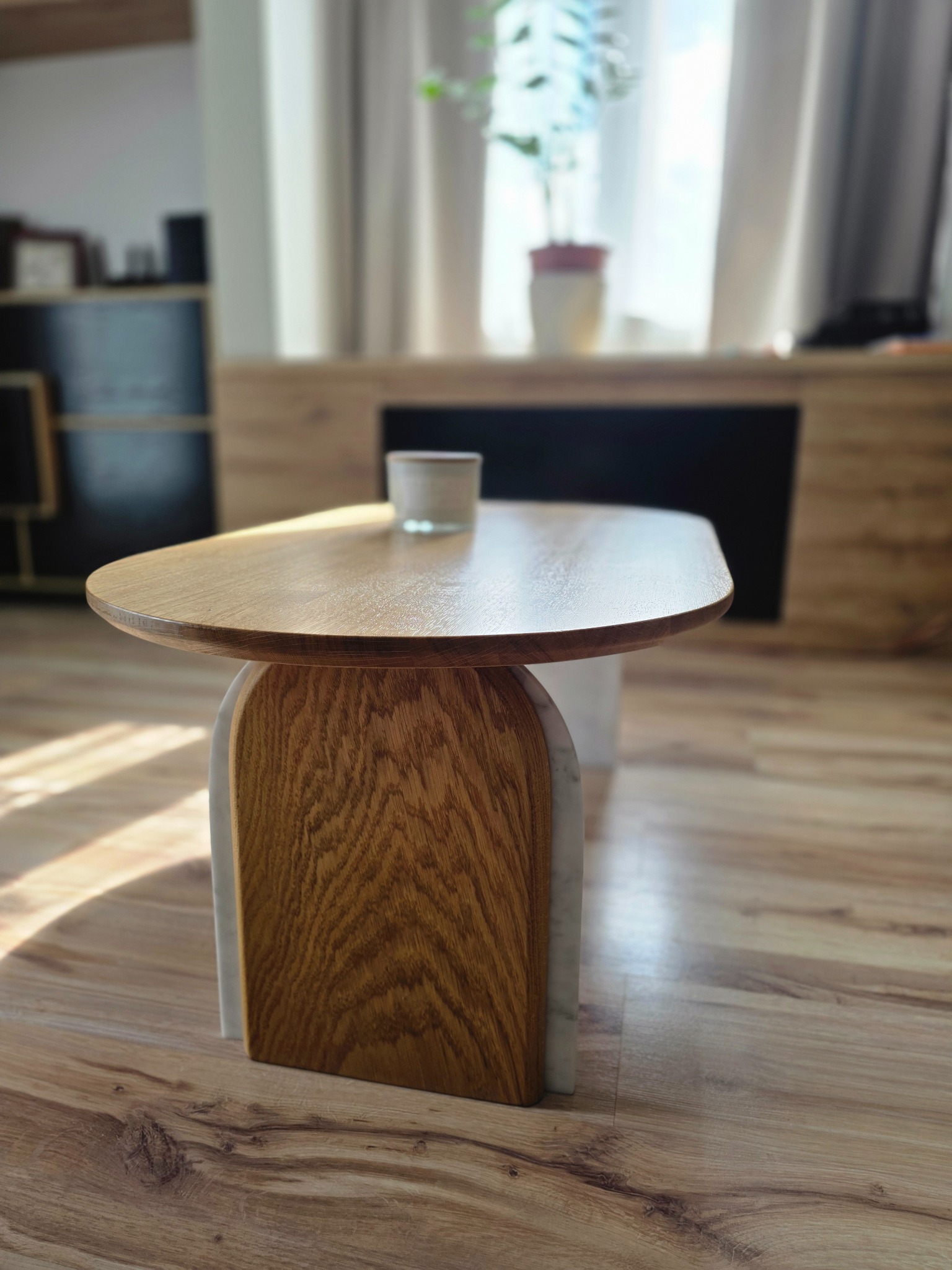 Stylowy stolik kawowy z blatem z jasnego drewna i oryginalną podstawą z drewnianych łuków z wstawkami z jasnego kamienia, na tle jasnego wnętrza.