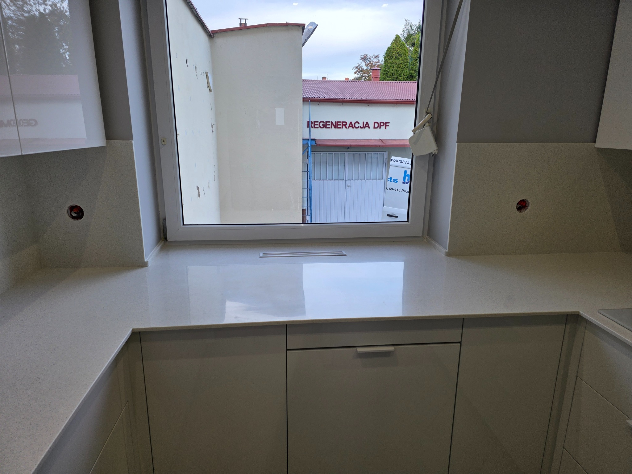 Jasny blat kuchenny z oknem, spod którego widać budynek warsztatu 'Regeneracja DPF'. Minimalistyczny design, neutralna kolorystyka. Widoczne szafki pod blatem.