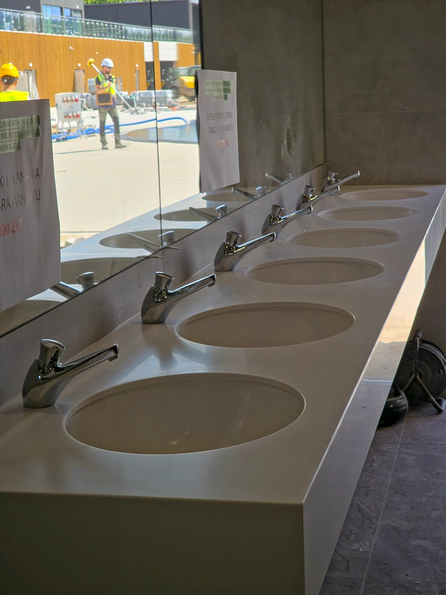 Długa, biała umywalka z kilkoma kranami, odbicie w lustrze pokazuje budowę na zewnątrz. Umywalka o minimalistycznym designie, z widocznymi znakami ostrzegawczymi.
