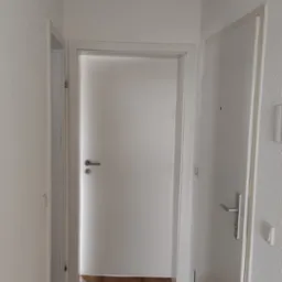 Jasne wnętrze korytarza z drewnopodobną podłogą, białymi ścianami i drzwiami. Widoczne detale wykończenia i minimalistyczny design.