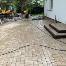 Ułożona beżowa kostka brukowa na podjeździe domu z jasną elewacją. W tle maszyna budowlana i materiały budowlane. Widoczne schody wejściowe z kostki w innym odcieniu.
