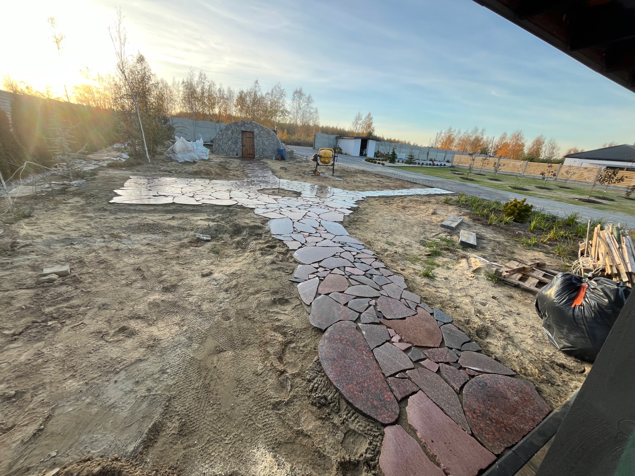 Kaskadowa ścieżka z nieregularnych płyt kamiennych w ogrodzie w trakcie budowy, z widoczną ziemią, mieszarką i domkiem narzędziowym w tle.