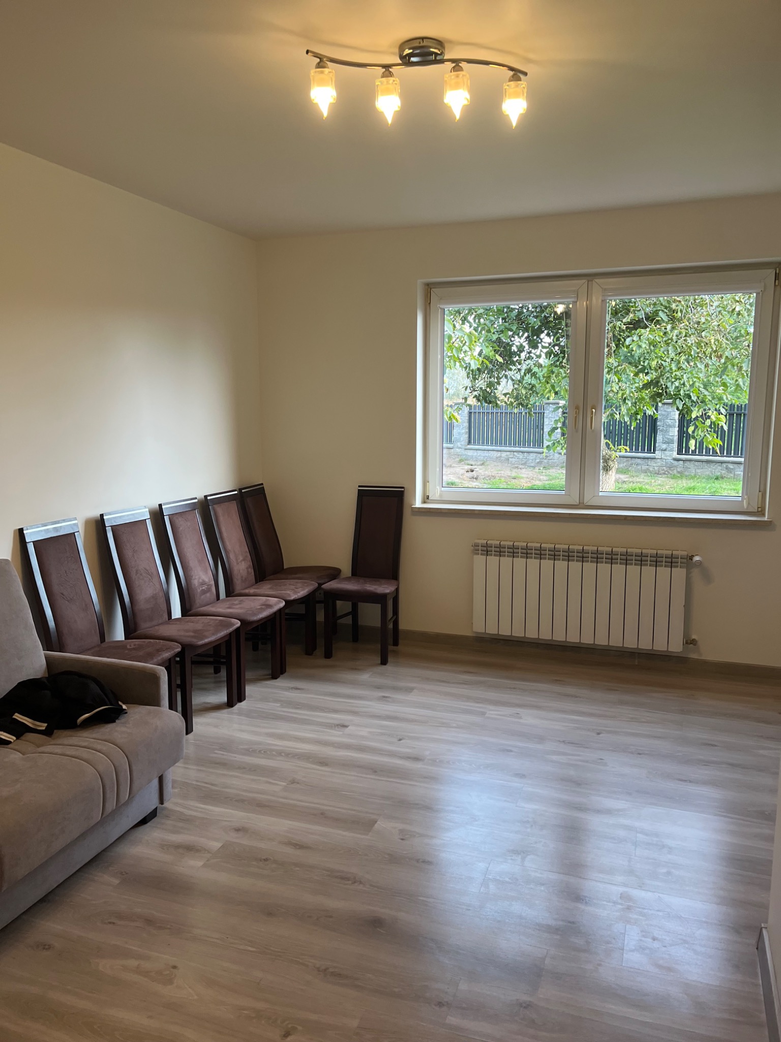 Jasne, przestronne wnętrze po malowaniu: kremowe ściany, drewnopodobna podłoga, rząd krzeseł, sofa, okno z widokiem na zieleń i biały grzejnik.