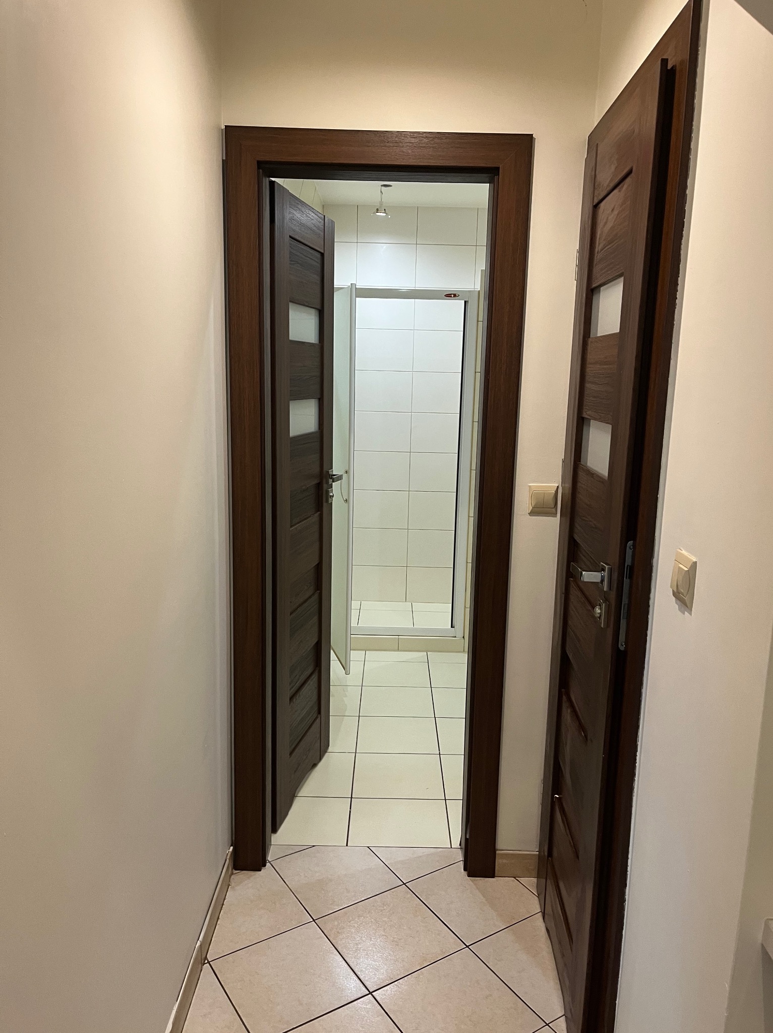 Wnętrze mieszkania po malowaniu: korytarz z dwiema otwartymi, drewnianymi drzwiami, widoczna biała łazienka w głębi. Jasne ściany, płytki podłogowe.