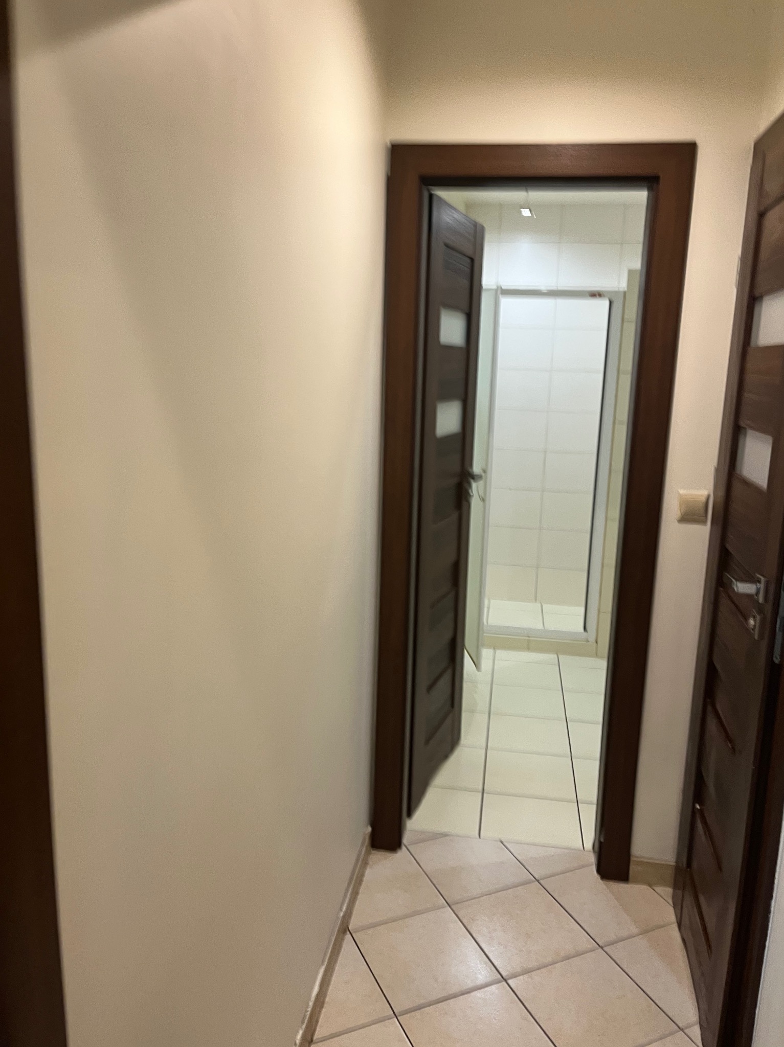 Otwarta drewniana, ciemna, wewnętrzna rama drzwiowa z widokiem na wykafelkowaną łazienkę z białą kabiną prysznicową, jasna ściana i podłoga z płytek.