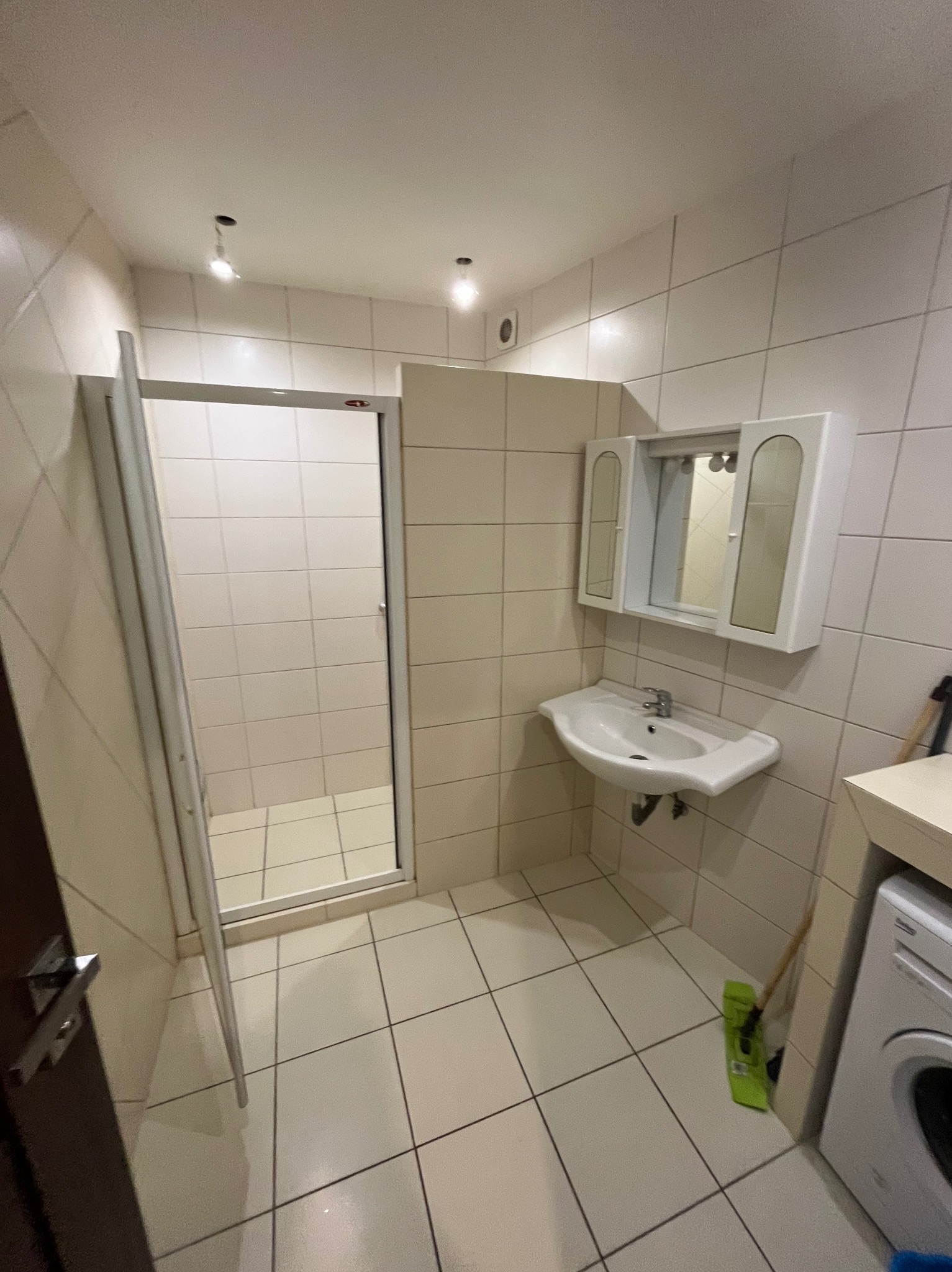 Jasna łazienka z kabiną prysznicową, umywalką z szafką i pralką. Widoczne białe płytki na ścianach i podłodze. Uwagę zwraca brak jednego elementu oświetlenia na suficie.