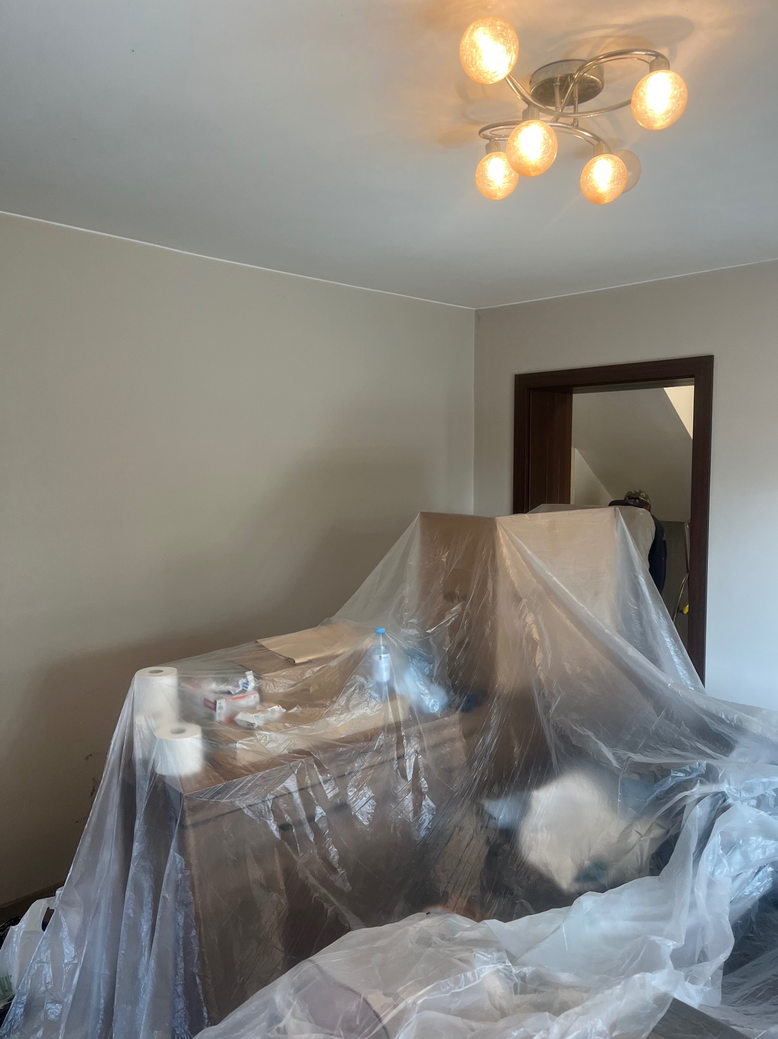 Pokój przygotowany do malowania: meble zakryte folią ochronną, ściany w jasnym odcieniu beżu, widoczna lampa sufitowa i fragment otwartych drzwi.