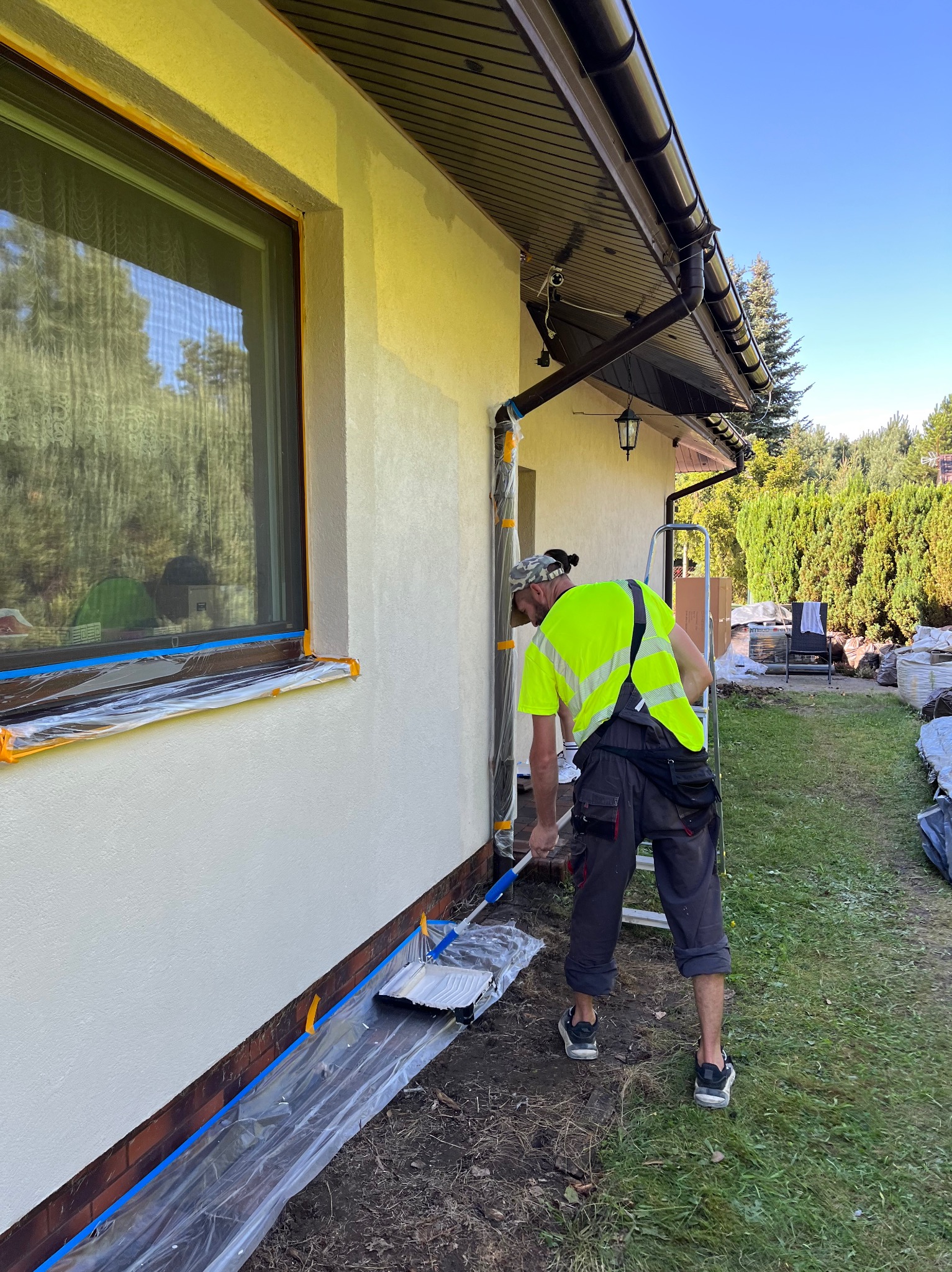 Malowanie elewacji domu w Łodzi. Mężczyzna maluje ścianę wałkiem, zabezpieczone okno i cokół. Widoczne narzędzia i drabina.
