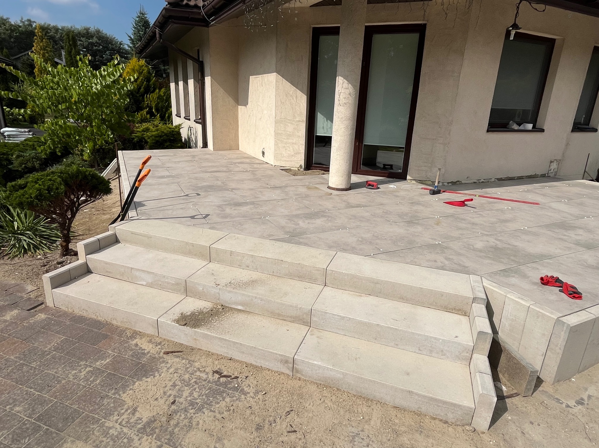 Jasnoszara, nowa nawierzchnia z płyt na tarasie domu jednorodzinnego. Widoczne schody z kostki i narzędzia brukarskie.