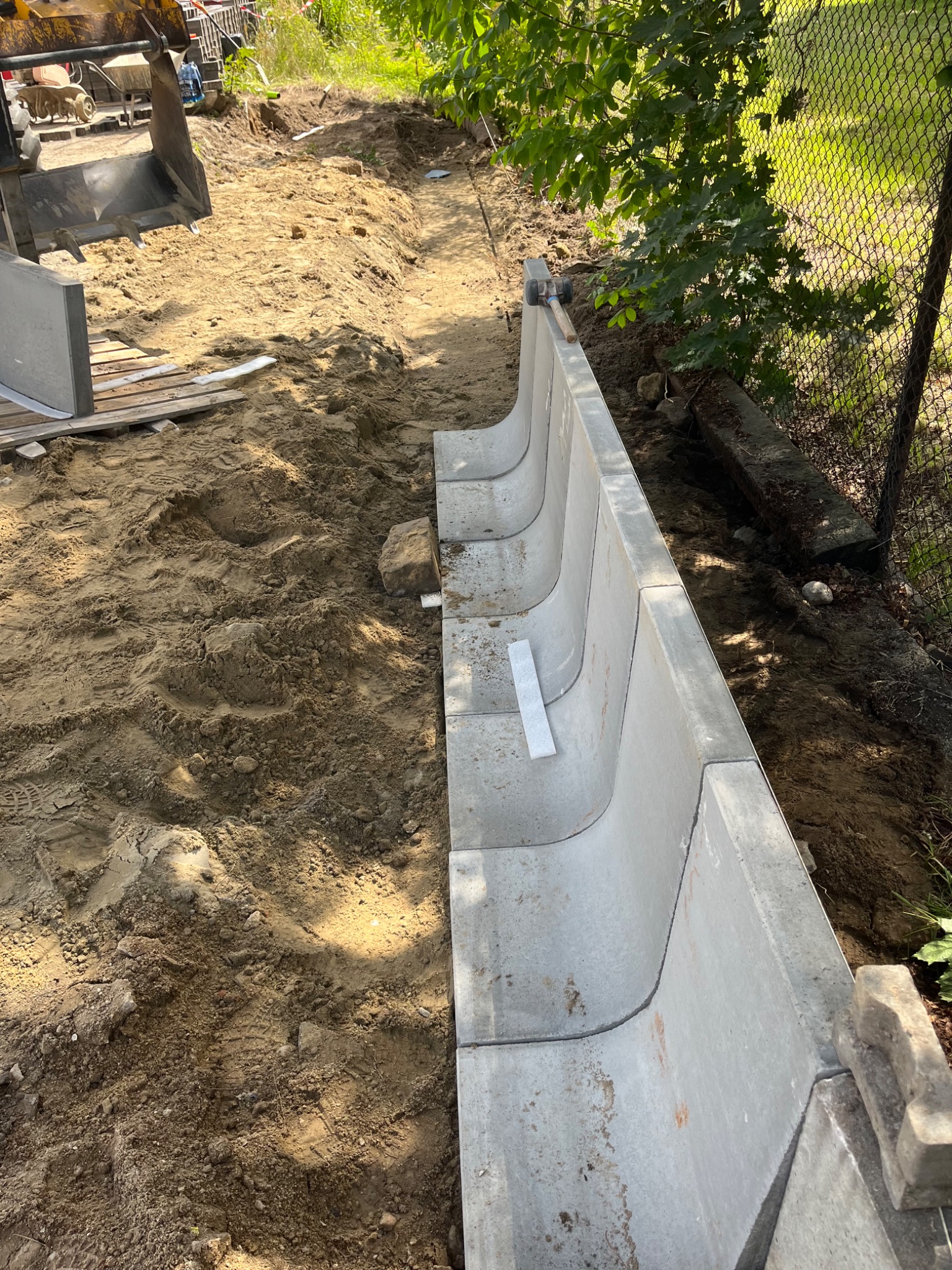 Montaż korytek odwadniających z betonu w ziemi, widoczny wykop i ciężki sprzęt. Korytka ułożone wzdłuż ogrodzenia z siatki, częściowo w cieniu drzew.