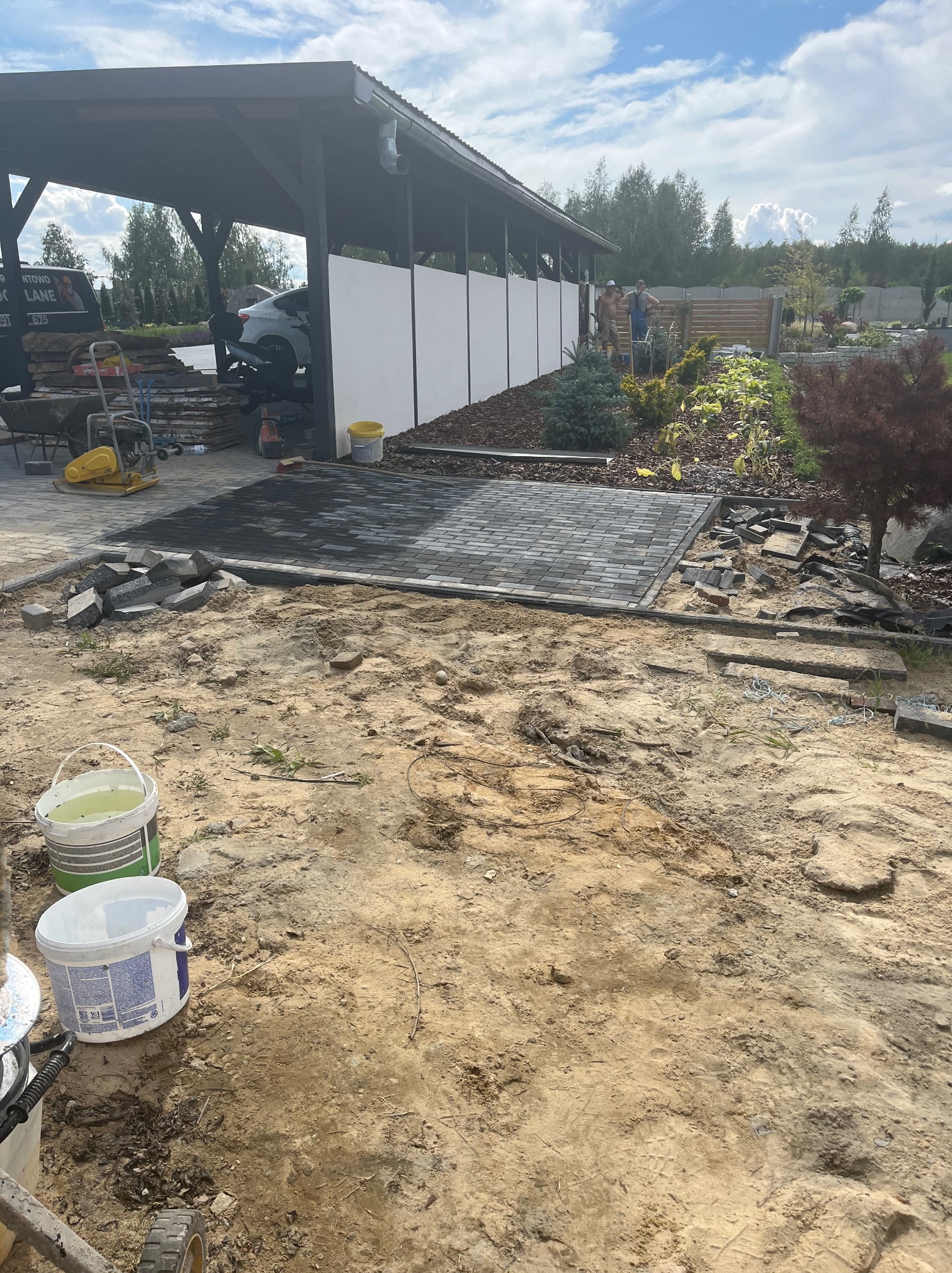 Fragment układanej kostki brukowej w odcieniach szarości na posesji z elementami ogrodu i zadaszeniem dla aut. Widoczne narzędzia i materiały budowlane.