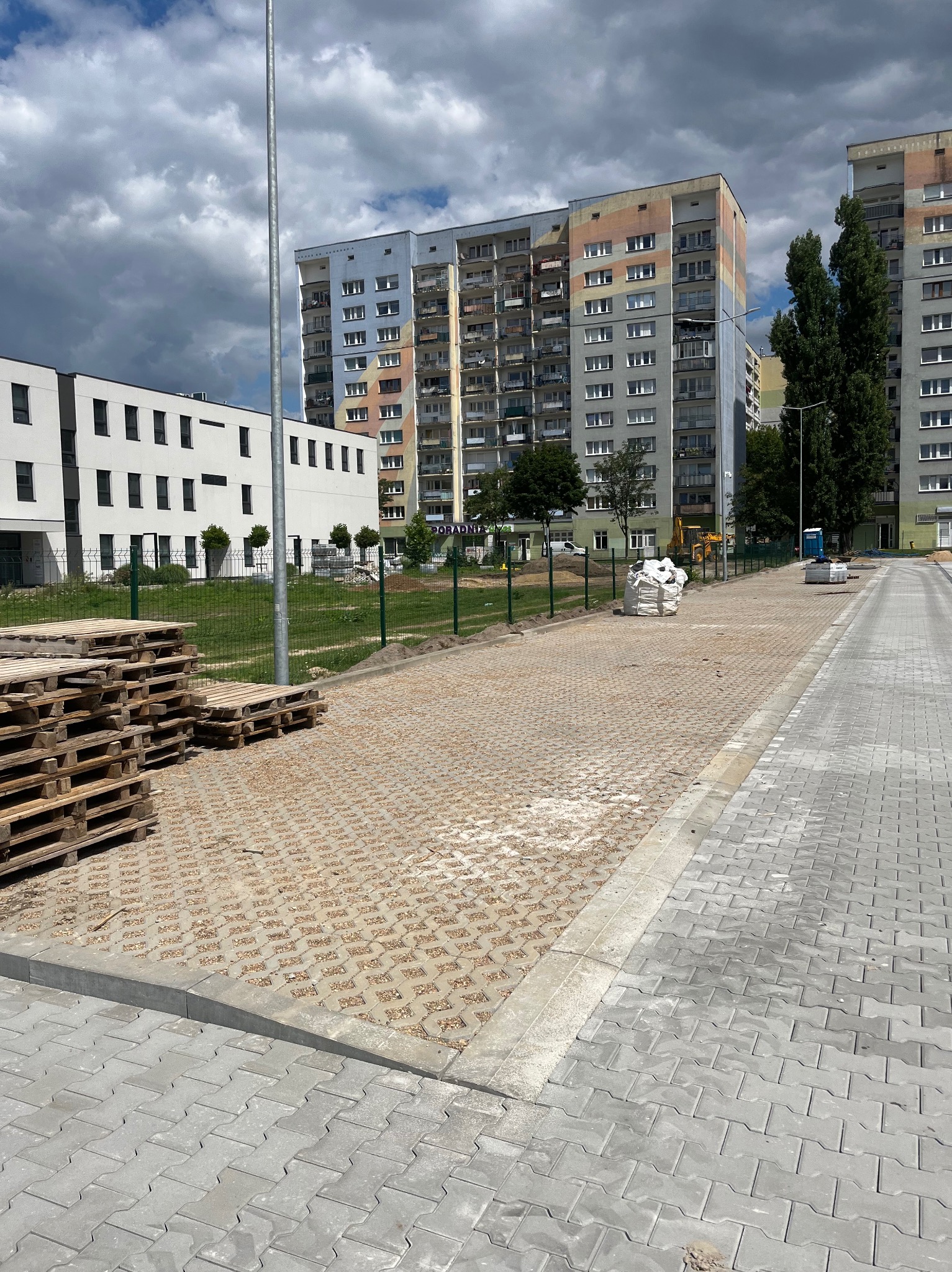 Częściowo ułożona, ażurowa kostka brukowa na parkingu osiedlowym z paletami i workami. W tle bloki mieszkalne i ogrodzenie.