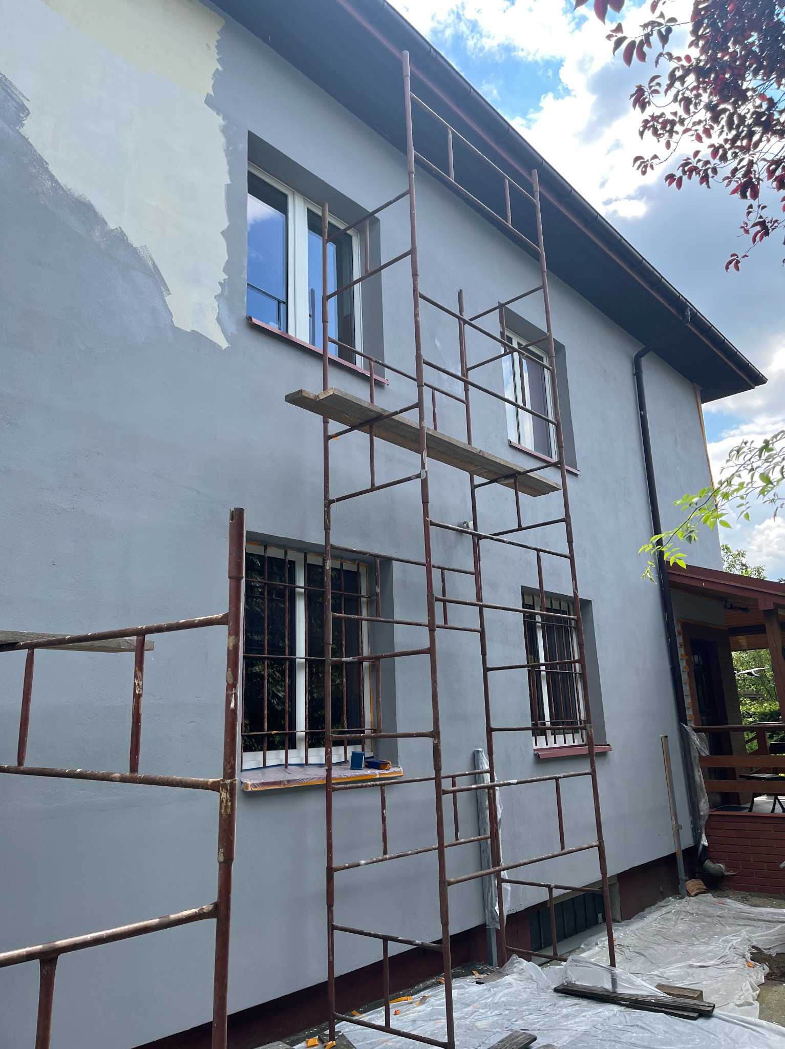 Szare, częściowo pomalowane ściany domu z rusztowaniem; widoczne okna z kratami. Etap prac malarskich z bliska, z folią ochronną na podłożu.