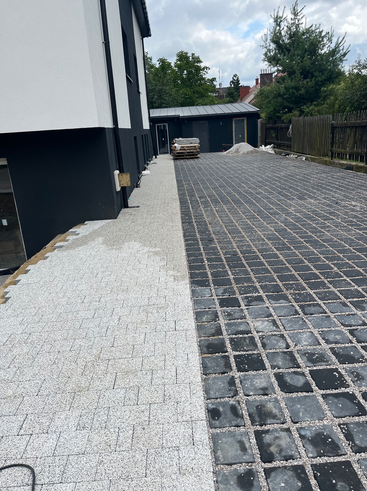 Częściowo ułożona, jasna kostka brukowa przechodzi w ciemną ażurową kostkę. W tle budynek i materiały budowlane. Widok z góry.