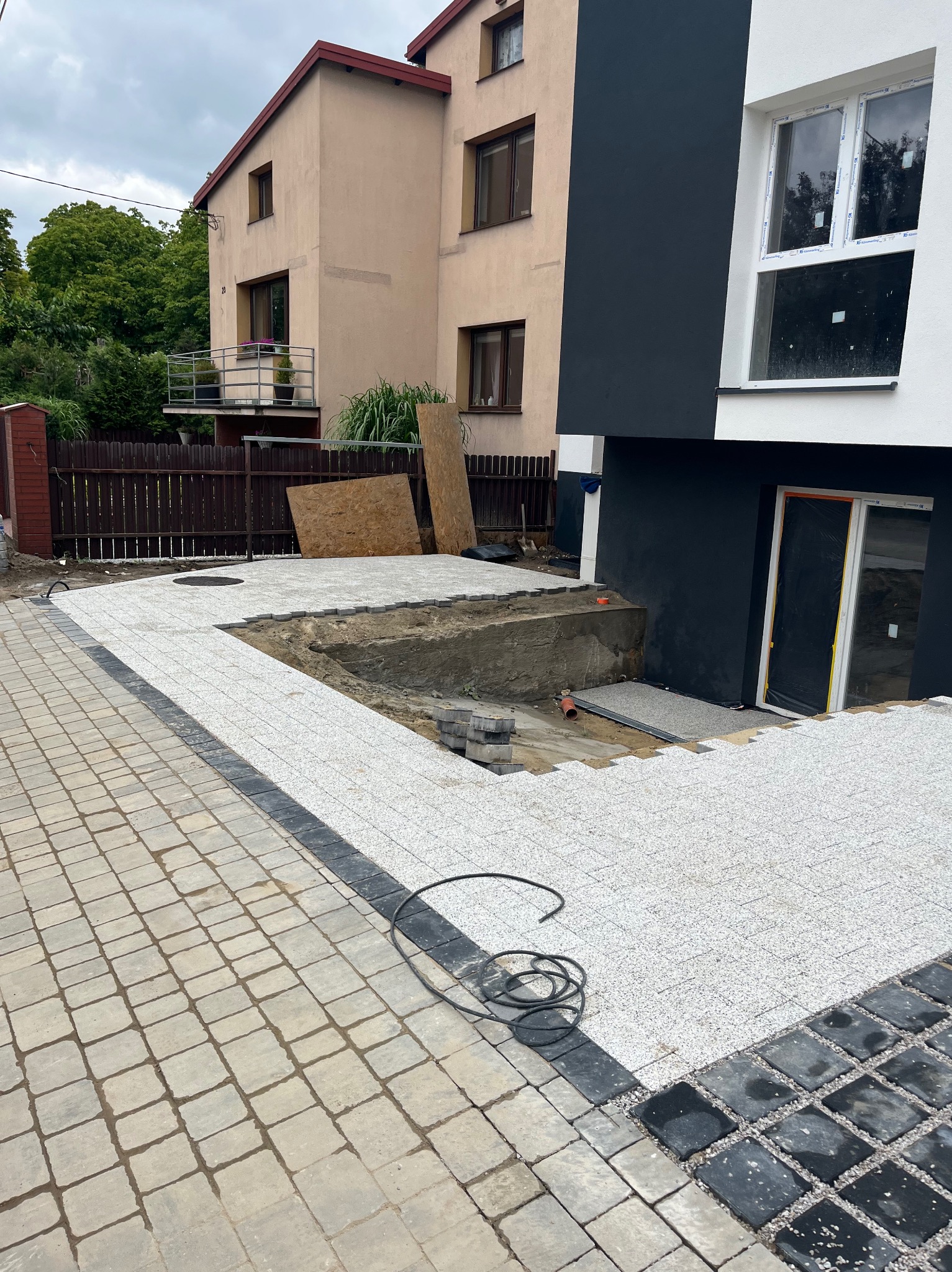 Fragment układania jasnoszarej kostki brukowej na posesji, widoczne różne etapy prac, tło stanowi budynek z czarno-białą elewacją i sąsiedni budynek.