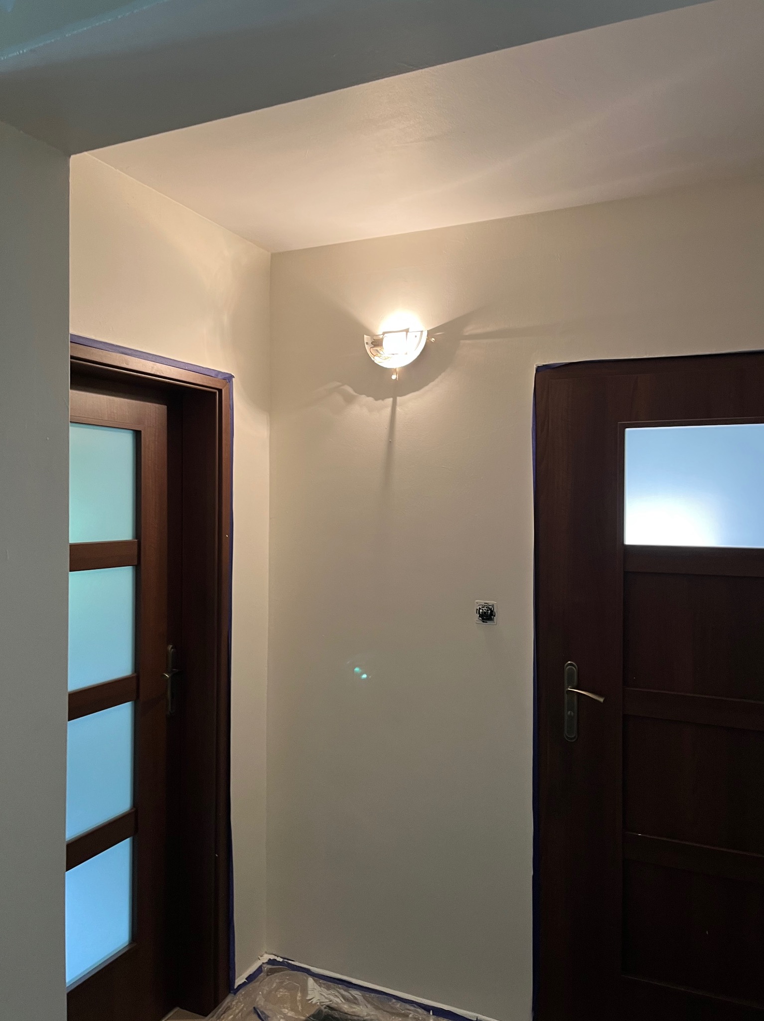 Świeżo pomalowany, jasny korytarz z dwoma ciemnymi drzwiami zabezpieczonymi taśmą malarską. Widoczna lampa ścienna i folia ochronna na podłodze.