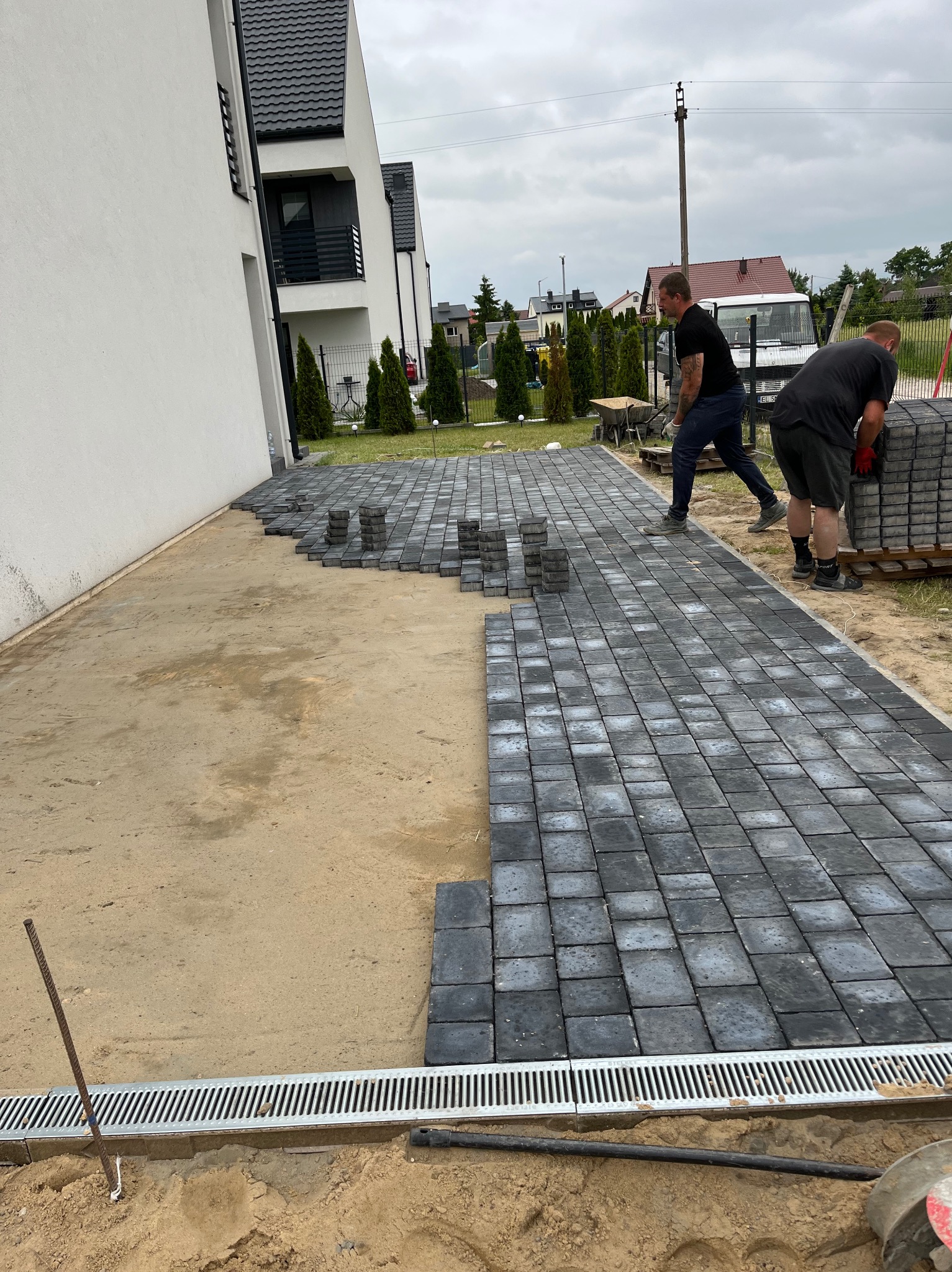 Układanie szarej kostki brukowej na posesji domu jednorodzinnego. Widoczny fragment ułożonej kostki, paleta z kostką i dwóch brukarzy w trakcie pracy. Odwodnienie liniowe.
