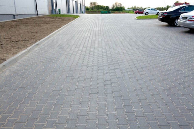 Szara kostka brukowa ułożona na parkingu przy budynku, z widocznymi samochodami i świeżo obsianą trawą. Krawężnik oddziela kostkę od trawnika.