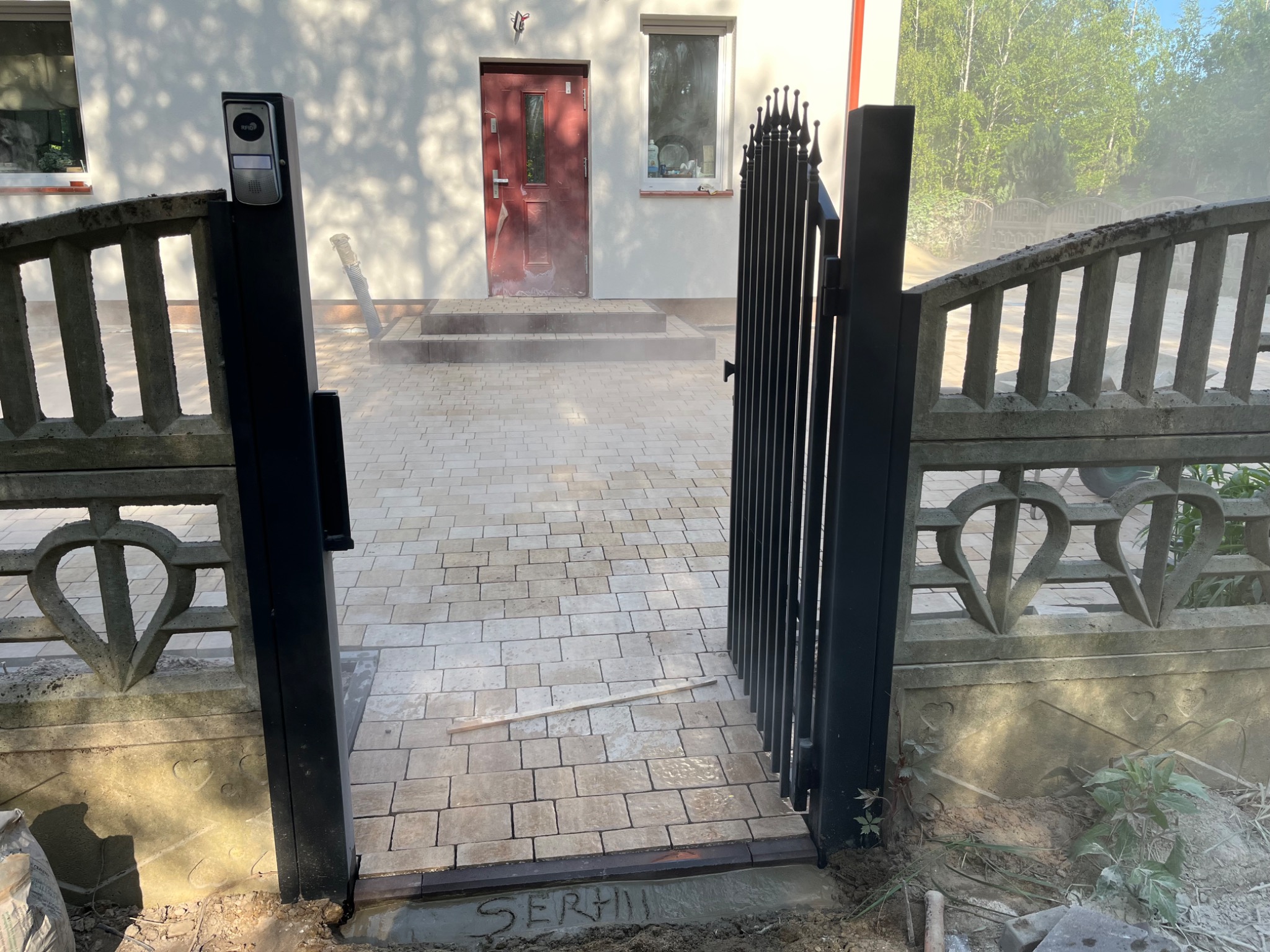 Ułożona kostka brukowa na podjeździe, widok przez otwartą, czarną bramę. W tle dom z czerwonymi drzwiami i betonowy płot z motywem serca.