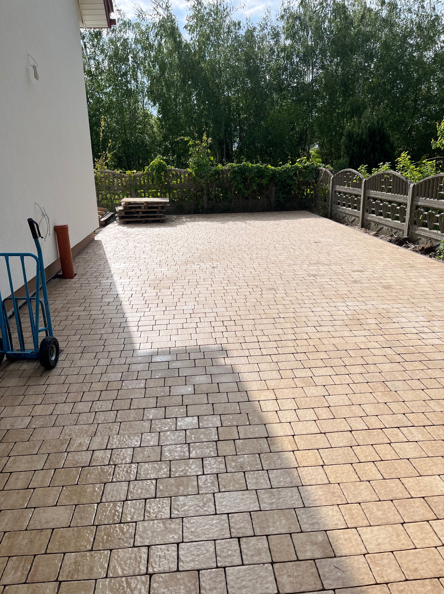 Ułożona, beżowa kostka brukowa na podjeździe, z widocznym wózkiem transportowym i paletami w tle, otoczona zielenią i ogrodzeniem, w słoneczny dzień.