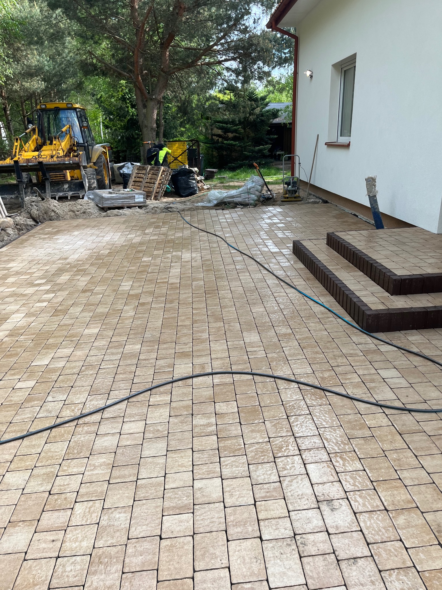 Ułożona beżowa kostka brukowa na podjeździe domu z jasną elewacją. W tle maszyna budowlana i materiały budowlane. Widoczne schody wejściowe z kostki w innym odcieniu.