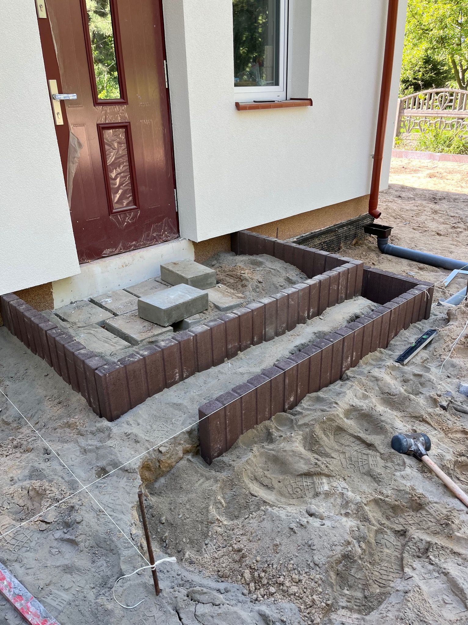 Montaż obrzeży schodów wejściowych z kostki. Widoczne narzędzia i poziomica. Prace brukarskie w toku, częściowo ułożona kostka. Szare obrzeża i piaskowe podłoże.