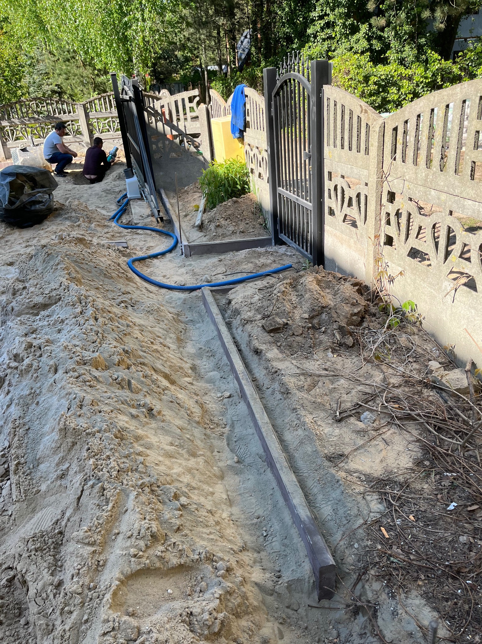 Przygotowanie podłoża pod kostkę brukową: wykopany rów z obrzeżami, widoczna brama i fragment ogrodzenia. Dwóch pracowników w tle.