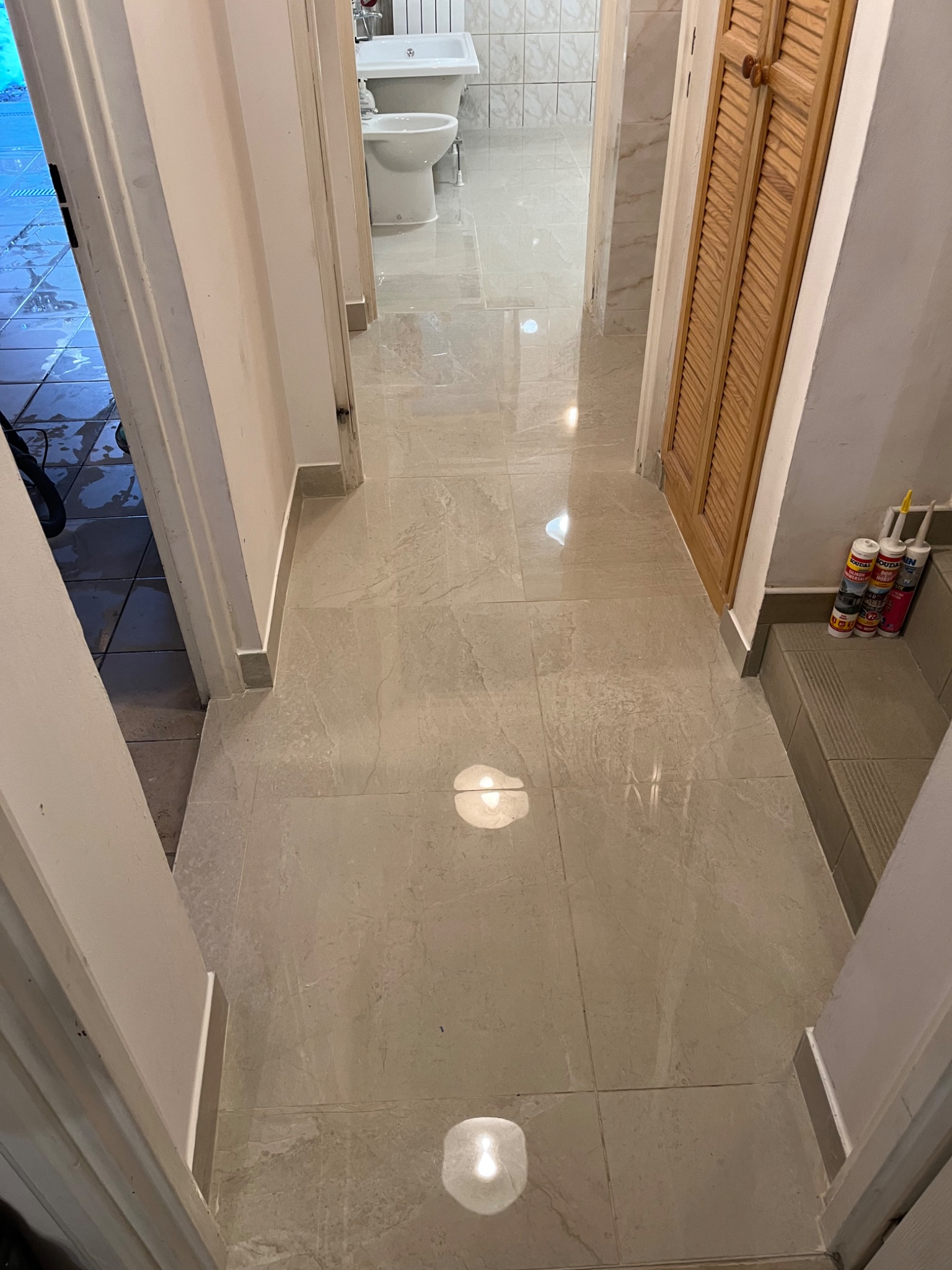 Wykończony korytarz z jasnymi, marmurkowymi płytkami podłogowymi, prowadzący do łazienki z białą armaturą. Widoczne tuby z silikonem przy ścianie.