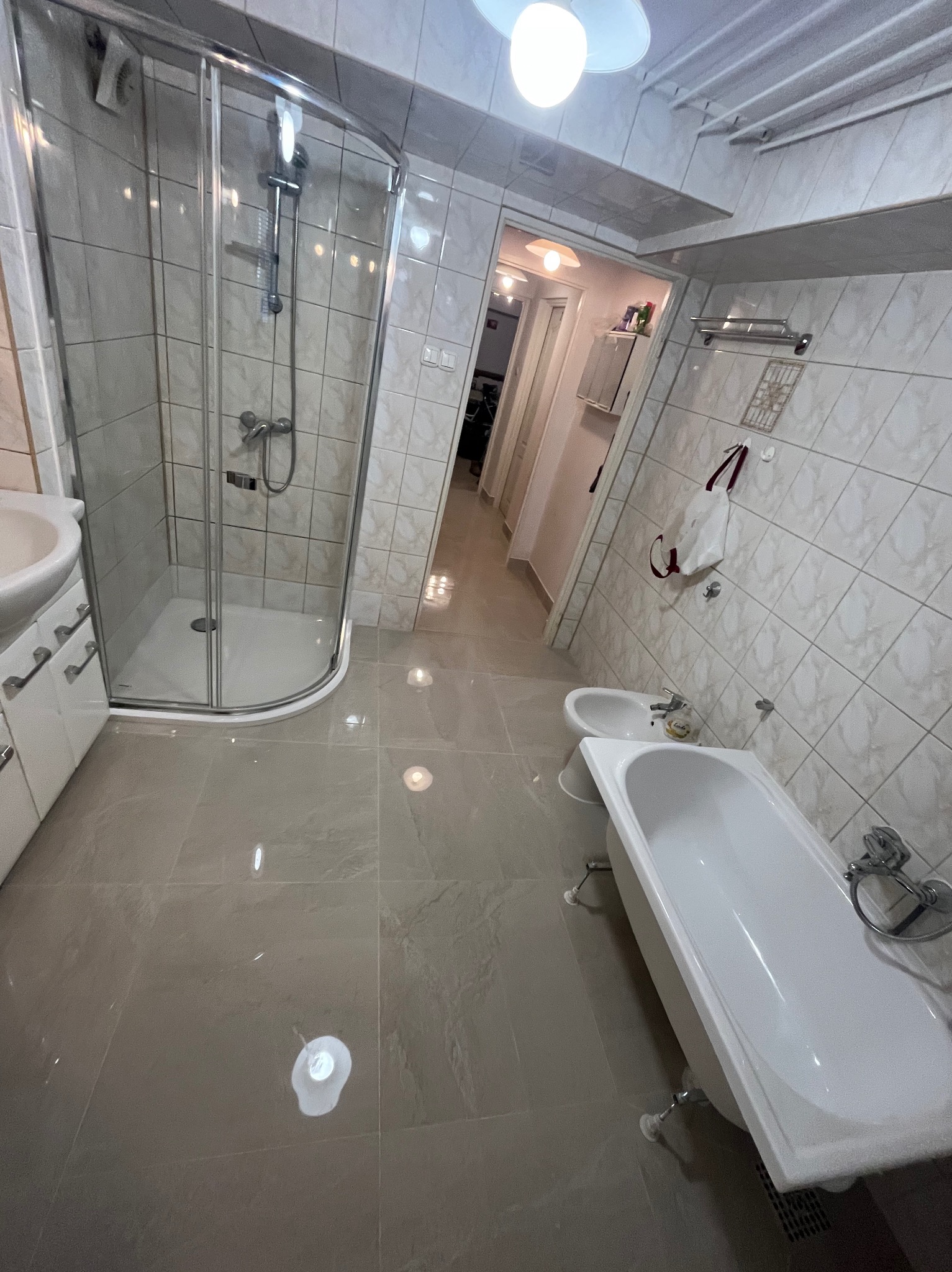 Wyremontowana łazienka z kabiną prysznicową, wanną na nóżkach i bidetem. Beżowe płytki na ścianach i podłodze. Widok na korytarz z oświetleniem.