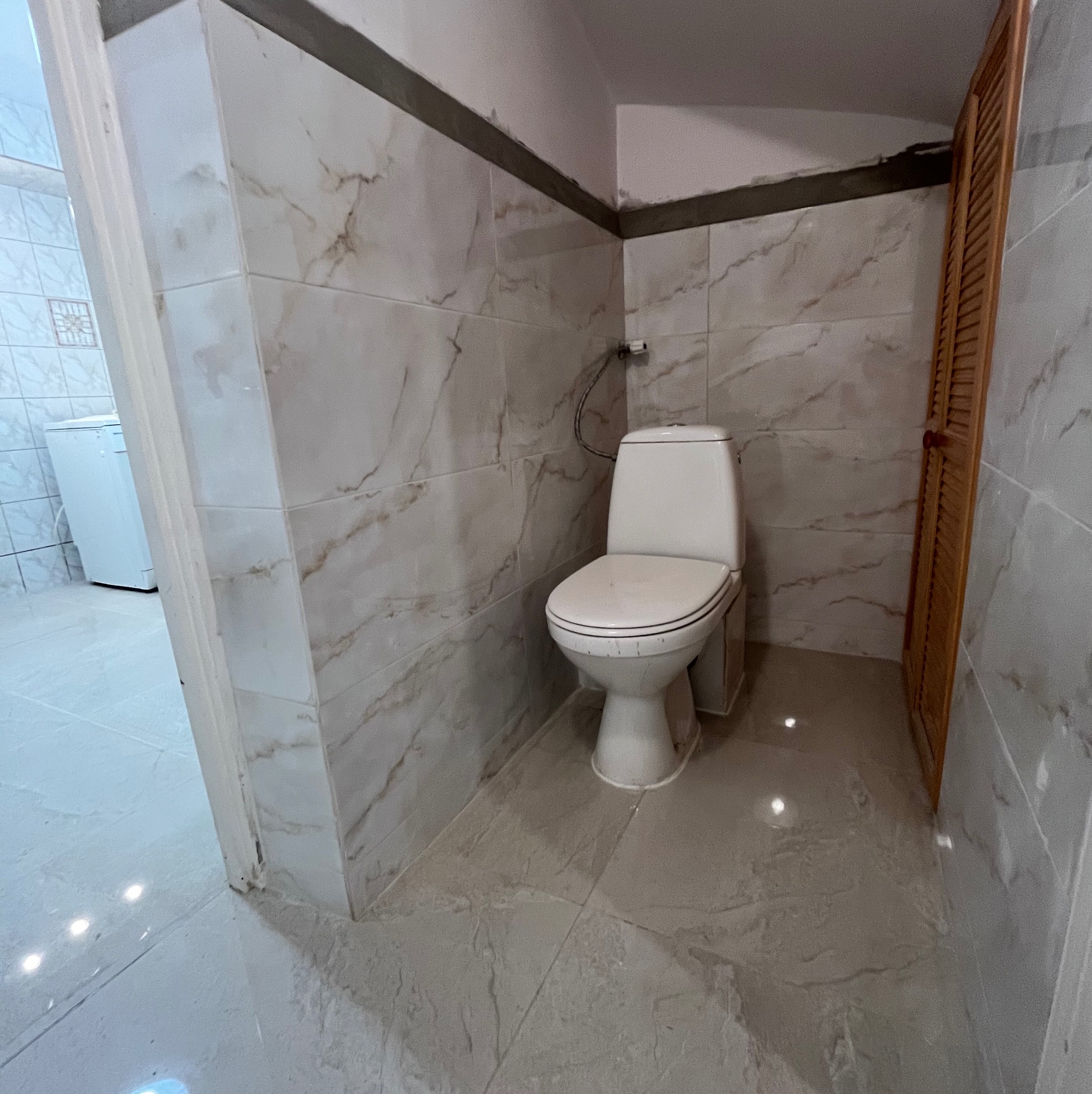 Wykończona łazienka z marmurowymi płytkami i nową toaletą. Widoczna drewniana szafka i pralka w tle. Ujęcie z perspektywy wejścia do pomieszczenia.