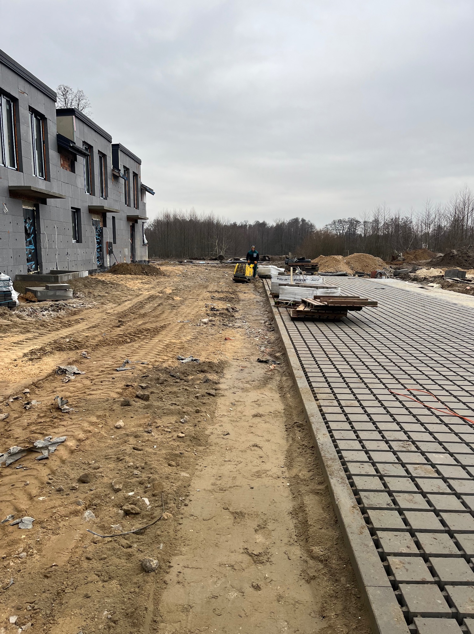 Układanie podbudowy pod kostkę brukową na terenie budowy osiedla. Widoczny mini-koparka i palety z kostką. Szara, pochmurna pogoda.