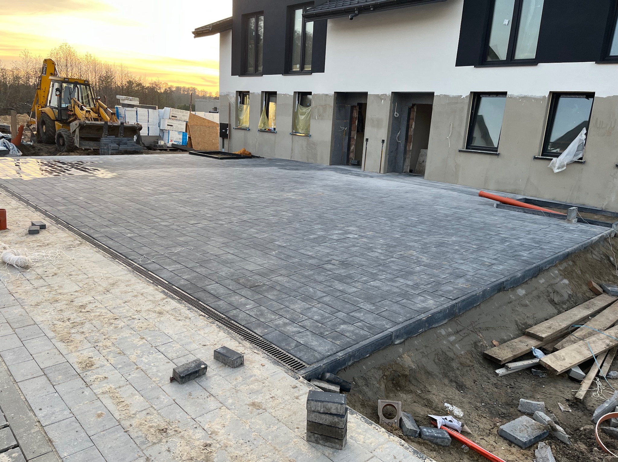 Ułożona, szara kostka brukowa przed budynkiem z czarnymi oknami. Część kostki jasna, jeszcze nie wyczyszczona. W tle koparka i zachodzące słońce.