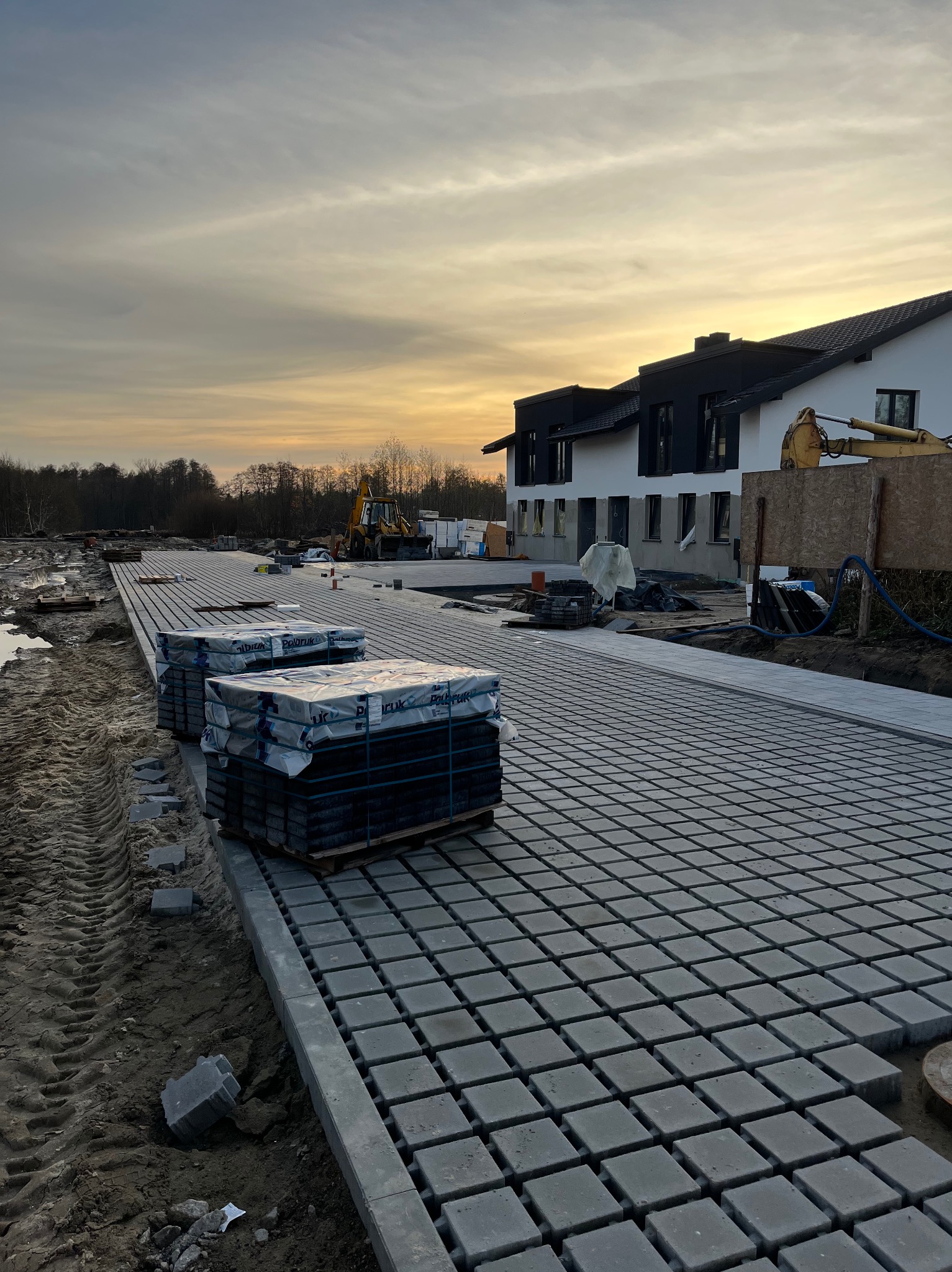 Układanie szarej kostki brukowej na posesji, palety z kostką w tle, widoczny budynek i koparka. Praca w toku, zachód słońca.