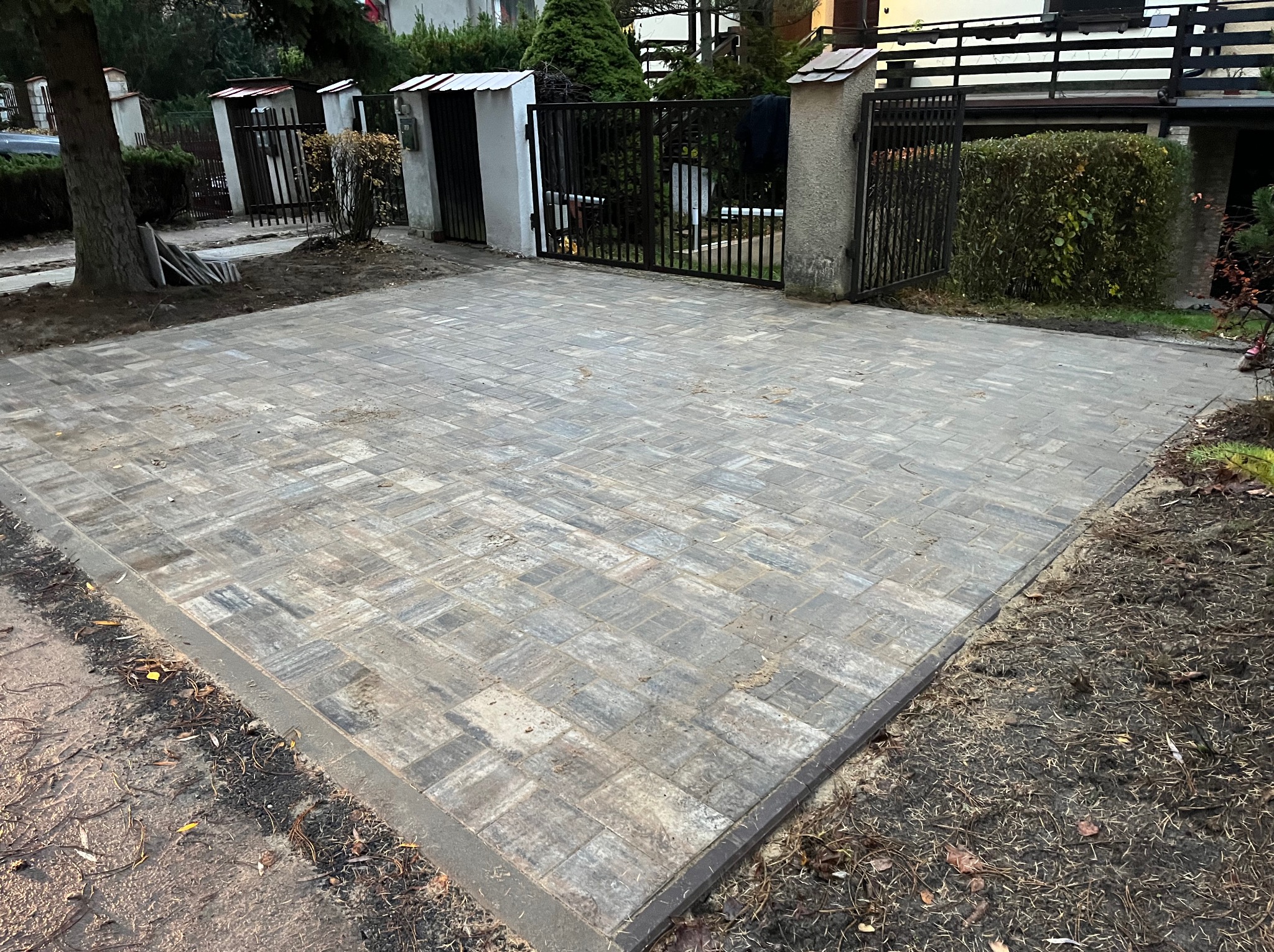 Ułożona kostka brukowa w odcieniach szarości tworzy wzór na podjeździe. Widoczne ogrodzenie posesji i drzewo. Całość w jesiennej scenerii.