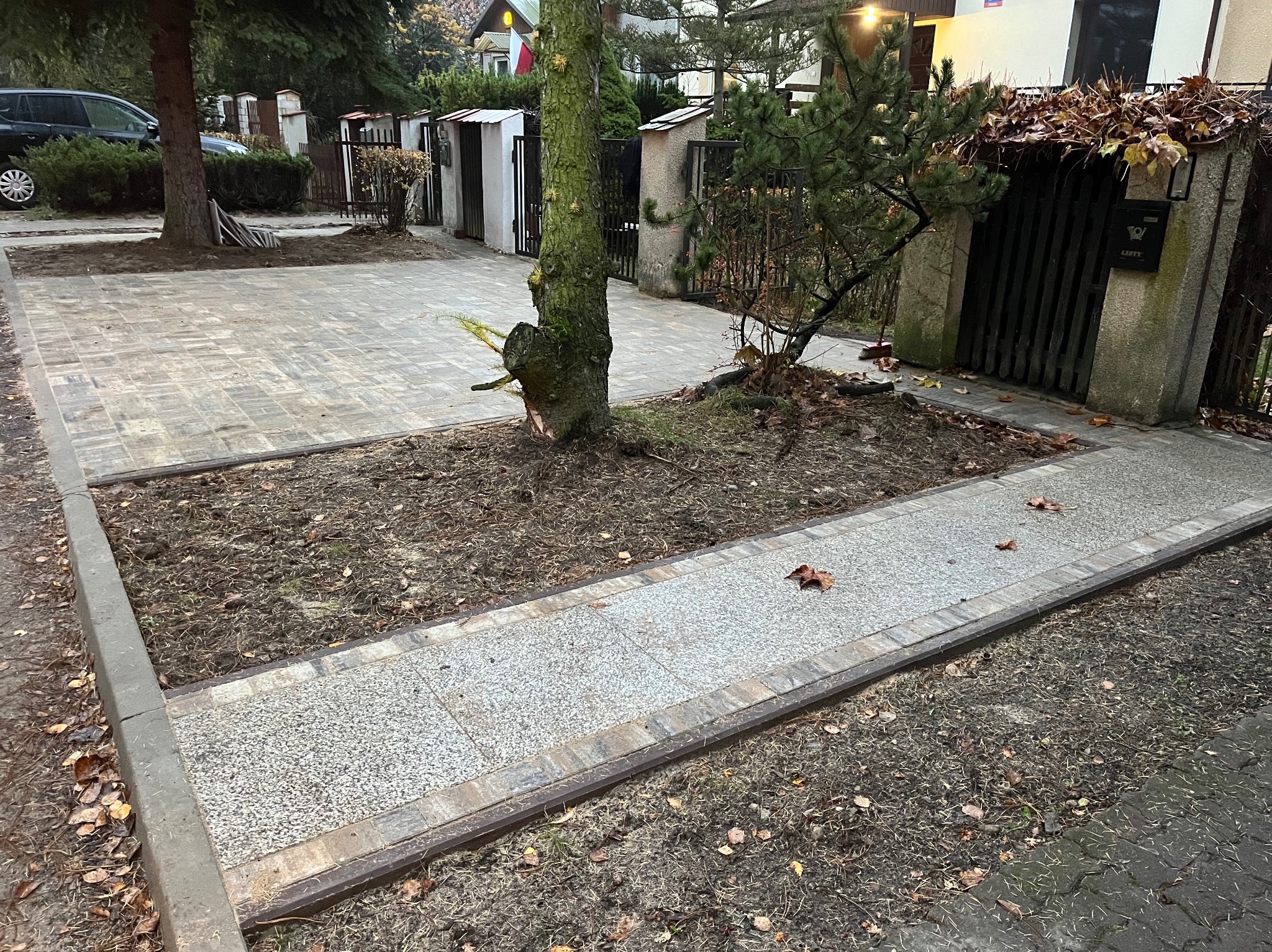 Ułożona kostka brukowa wokół drzewa na posesji, obrzeża z kostki, widok na furtkę i dom w tle, jesienne liście na ziemi.