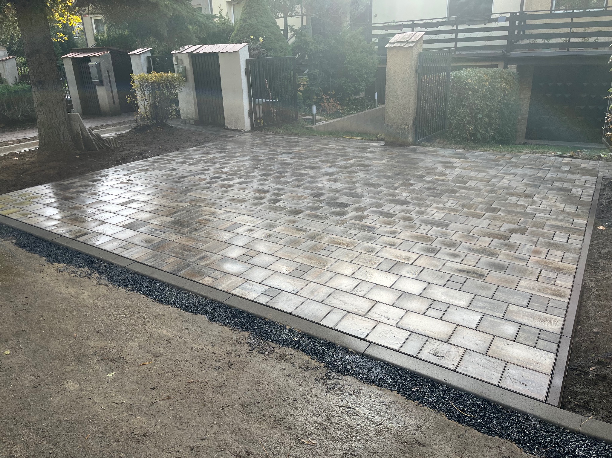 Ułożona kostka brukowa w odcieniach szarości i beżu, tworząca wzór na podjeździe. Widoczne obrzeża z drobnego kamienia i elementy architektury ogrodowej w tle.