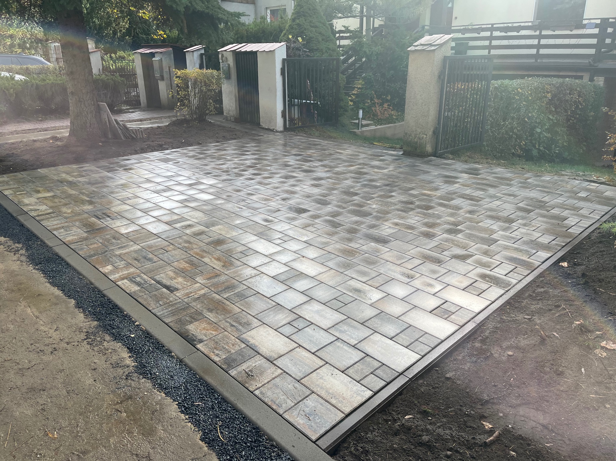 Ułożona kostka brukowa w Łodzi, wzór prostokątny z obramowaniem. Widok na posesję z bramą i zielenią w tle. Świeżo wykonana nawierzchnia.