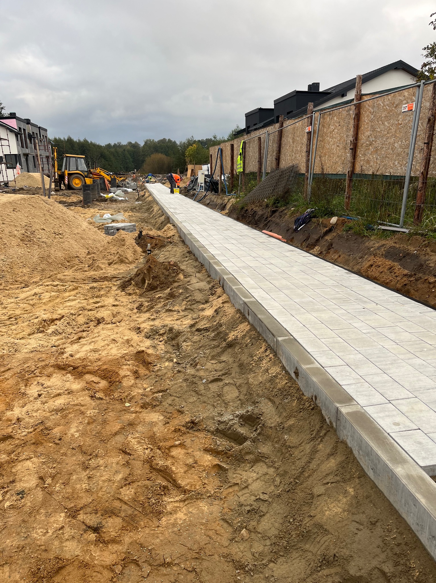 Długa, jasnoszara ścieżka z kostki brukowej z betonowymi obrzeżami, w trakcie budowy. W tle koparka i budynki w stanie surowym, pochmurne niebo.