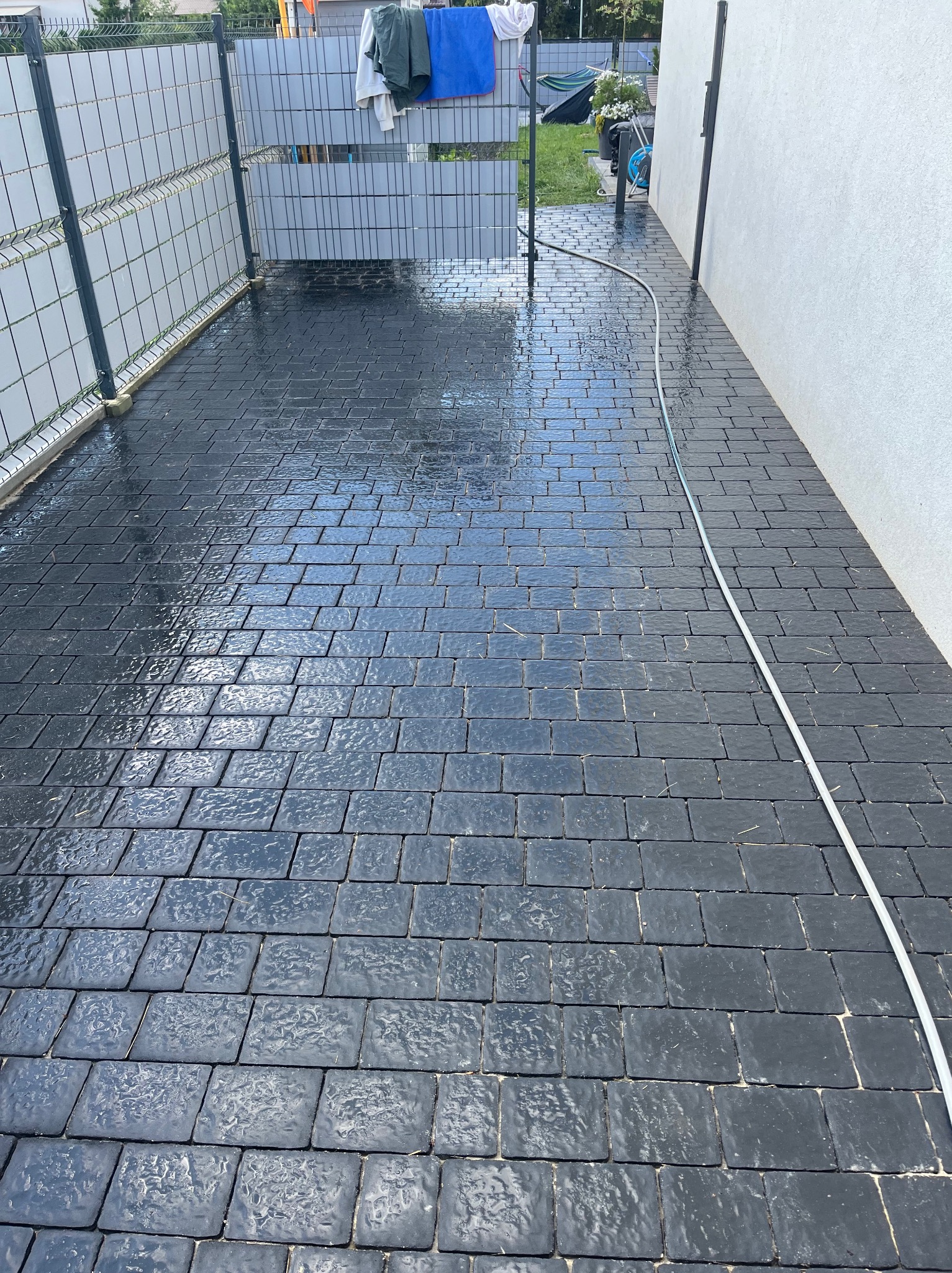 Umyta, ciemna kostka brukowa na podjeździe, mokra od wody, z widocznym wężem ogrodowym. W tle szary płot i fragment budynku z białymi ścianami.