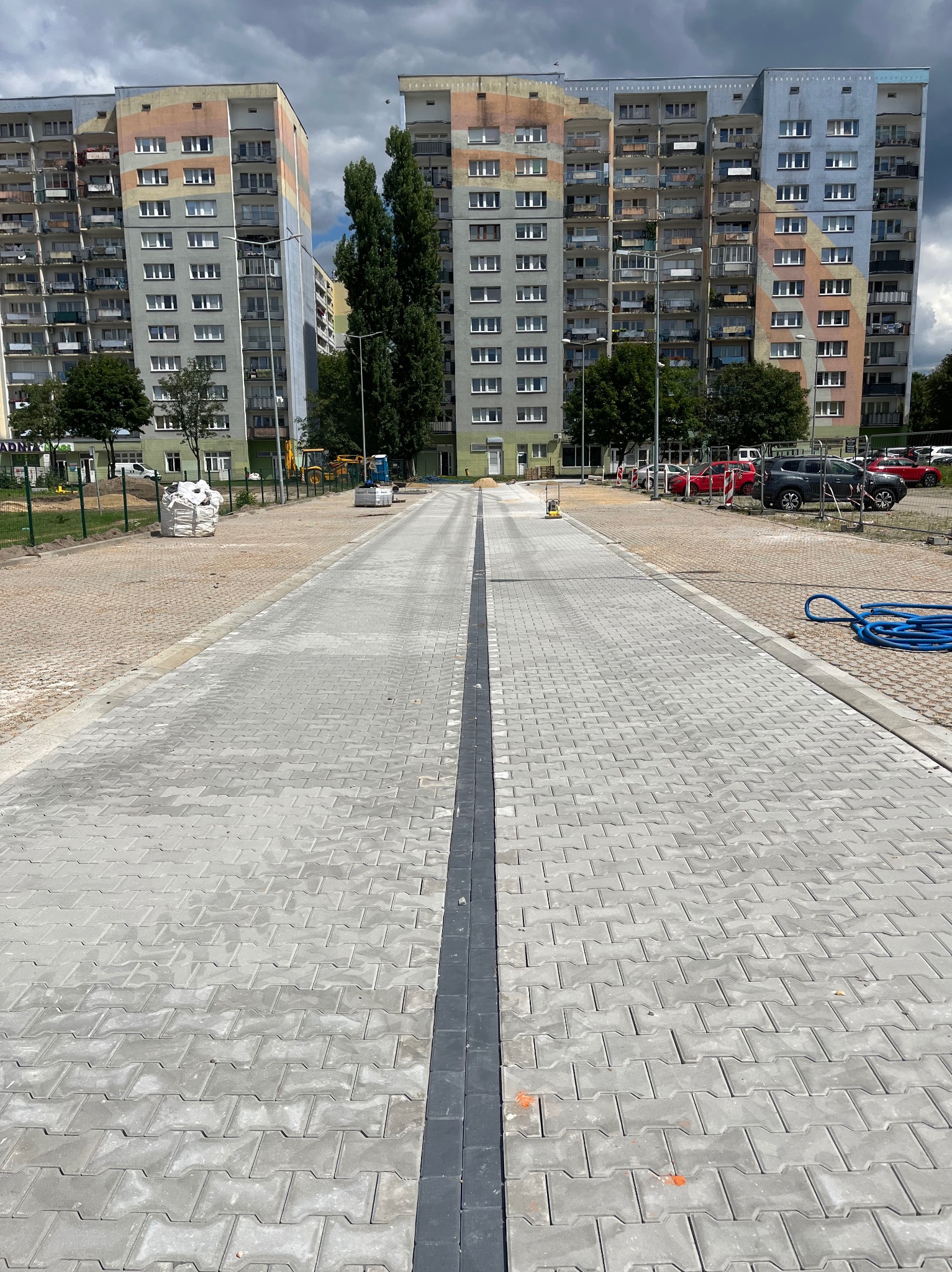 Nowo ułożona, szara kostka brukowa z ciemnoszarym pasem pośrodku, prowadząca w kierunku bloków mieszkalnych pod pochmurnym niebem. Widoczny sprzęt budowlany i zaparkowane samochody.