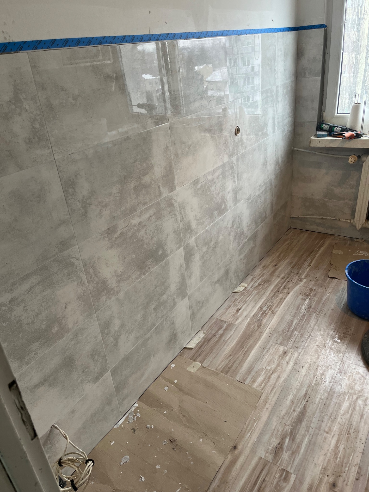 Ściana łazienki z szarymi, błyszczącymi płytkami, zabezpieczona niebieską taśmą. Podłoga częściowo wyłożona panelami, widoczne narzędzia i wiadro.