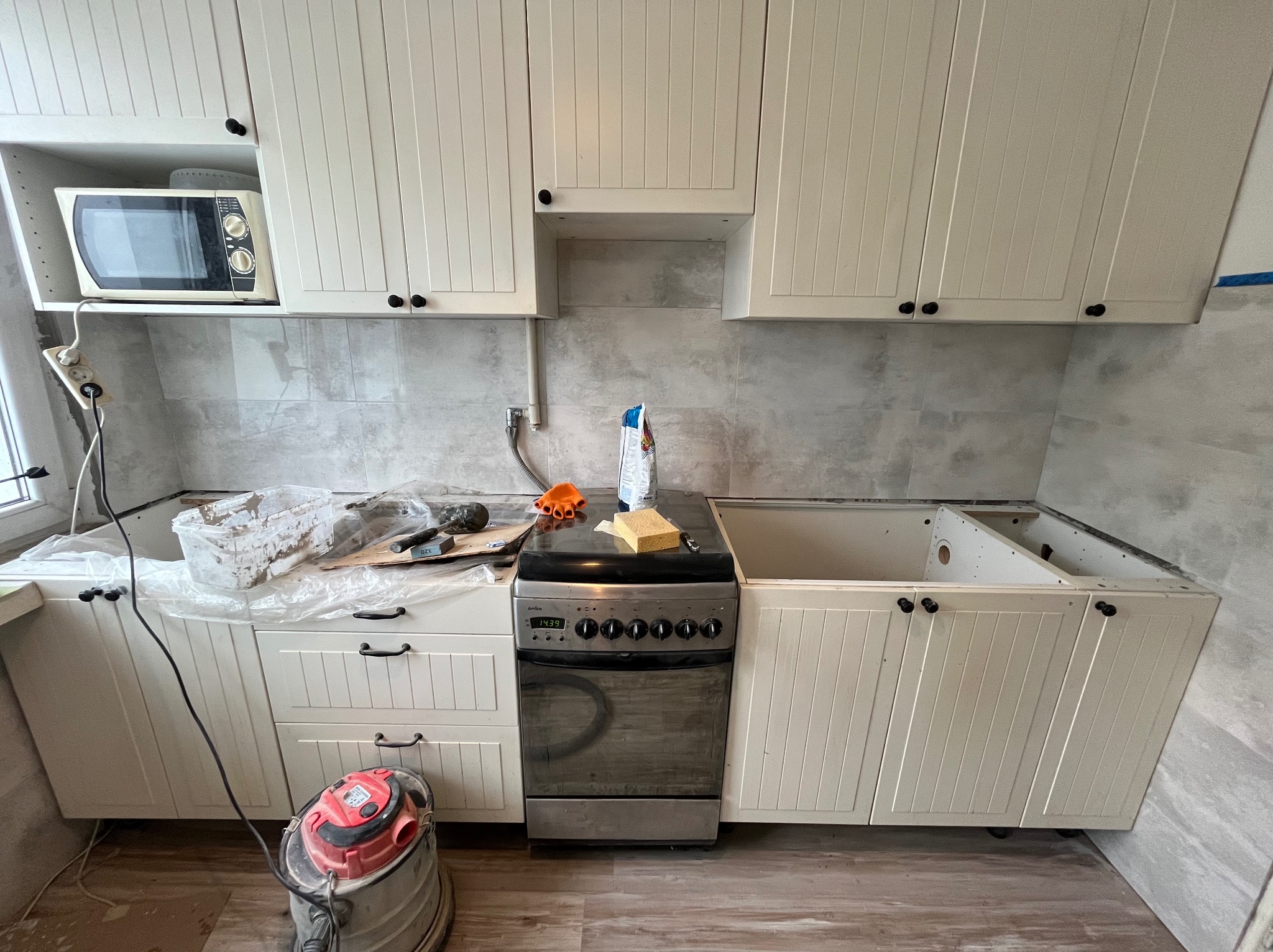 Montaż białych szafek kuchennych z frezowanymi frontami i czarnymi uchwytami. Widoczny fragment kuchenki i odkurzacz. Szare płytki na ścianie.