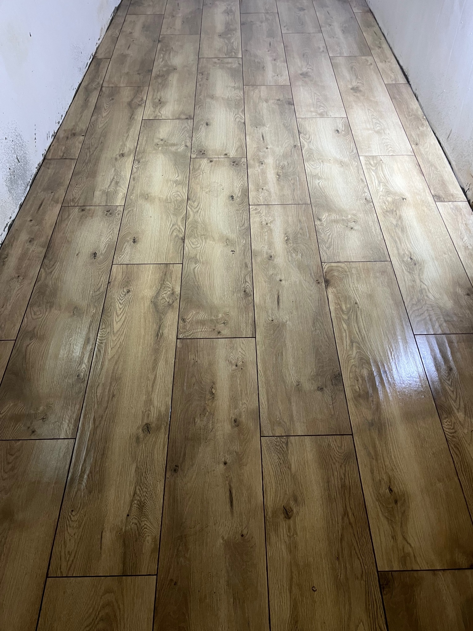 Ułożone panele podłogowe w wąskim pomieszczeniu, imitujące drewno, z widocznymi refleksami światła i niedomalowaną ścianą w tle.